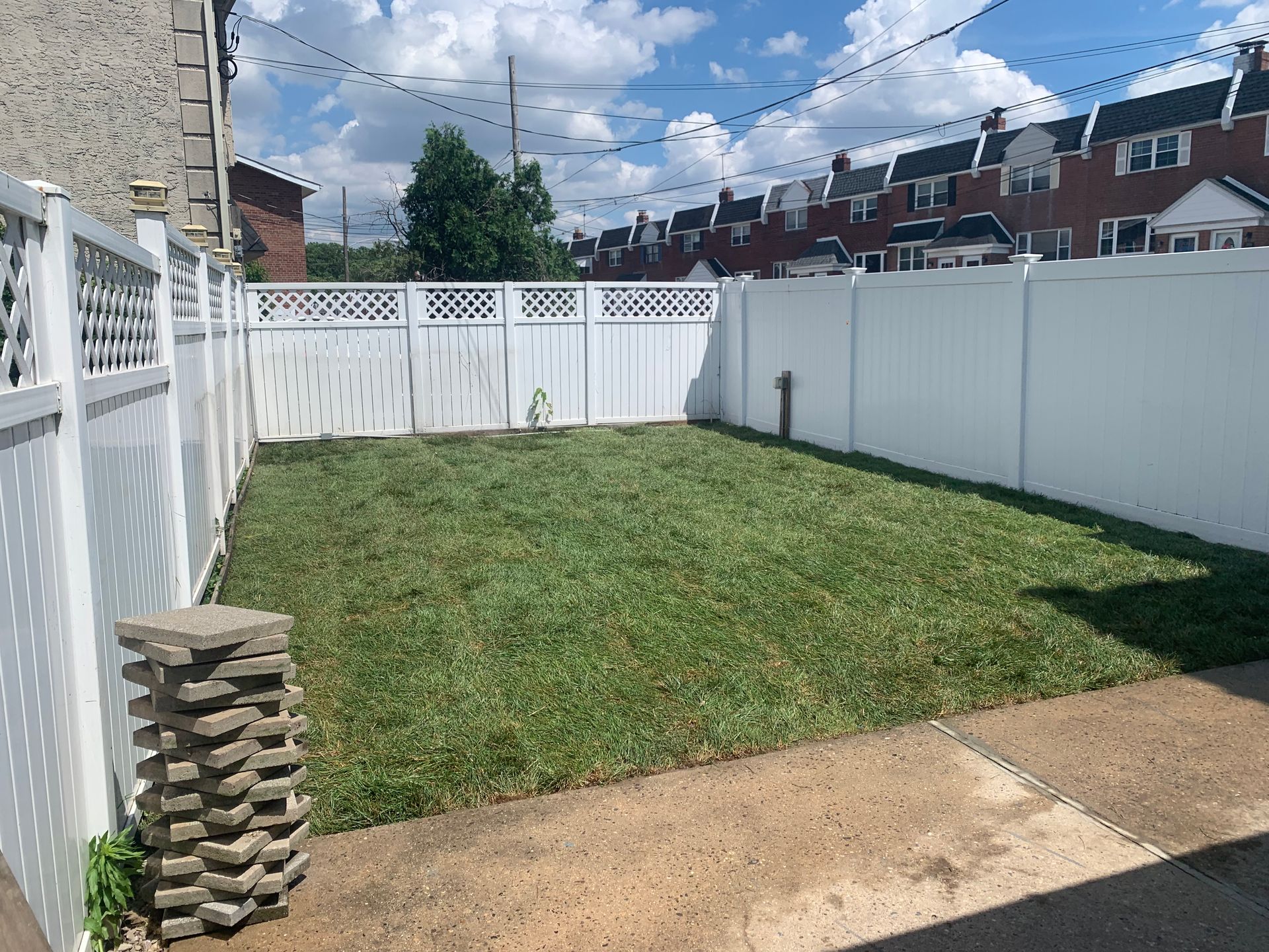 A backyard with a small, freshly cut green lawn enclosed by a white vinyl privacy fence under a blue cloudy sky.