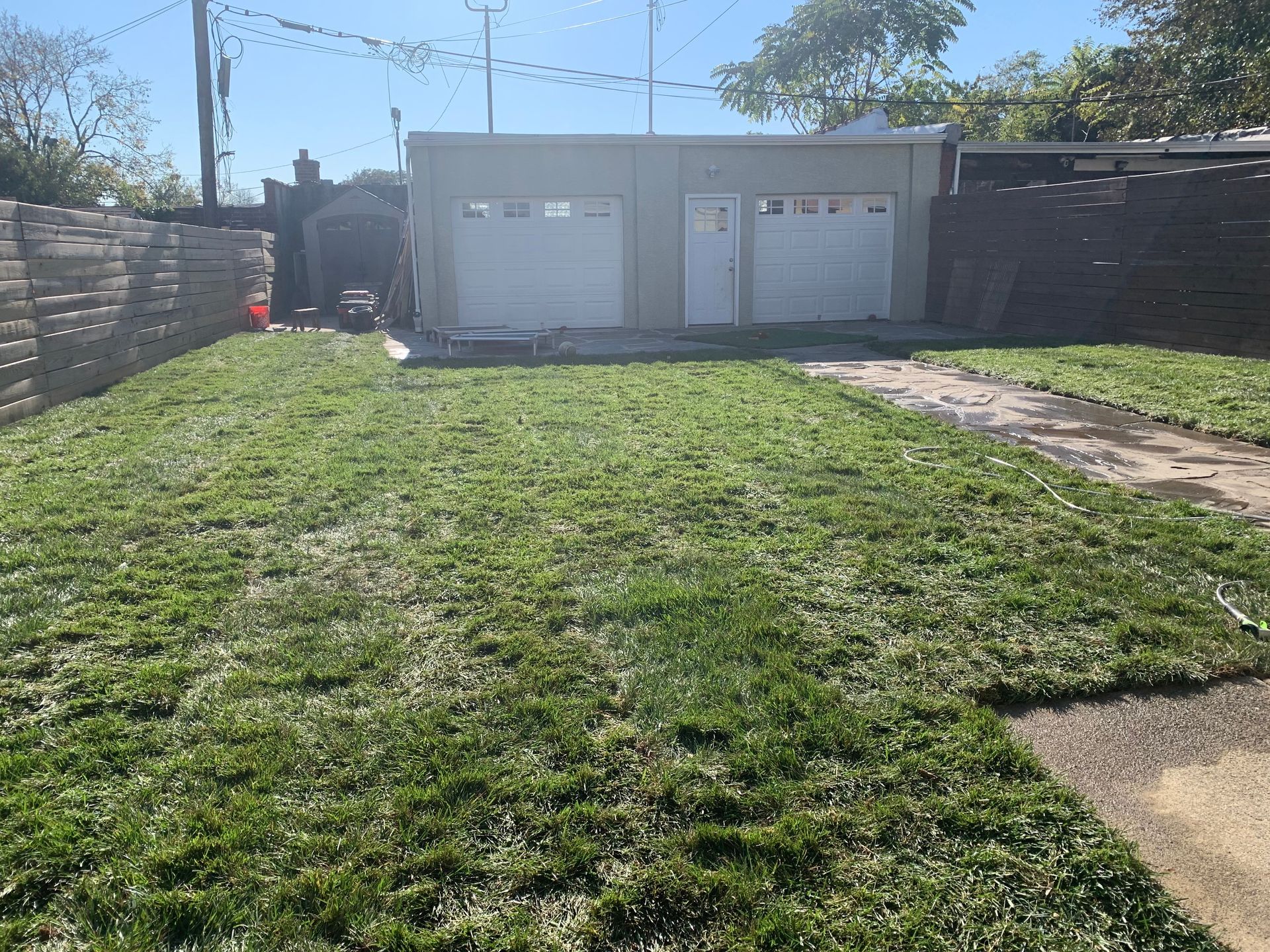 A backyard with a recently mowed lawn leading to a detached two-car garage with white doors under a clear blue sky.