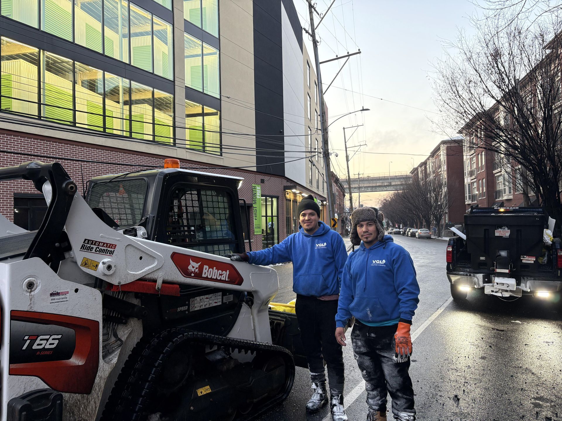 Two workers stand beside a Bobcat track loader on a city street. One points at the machine.