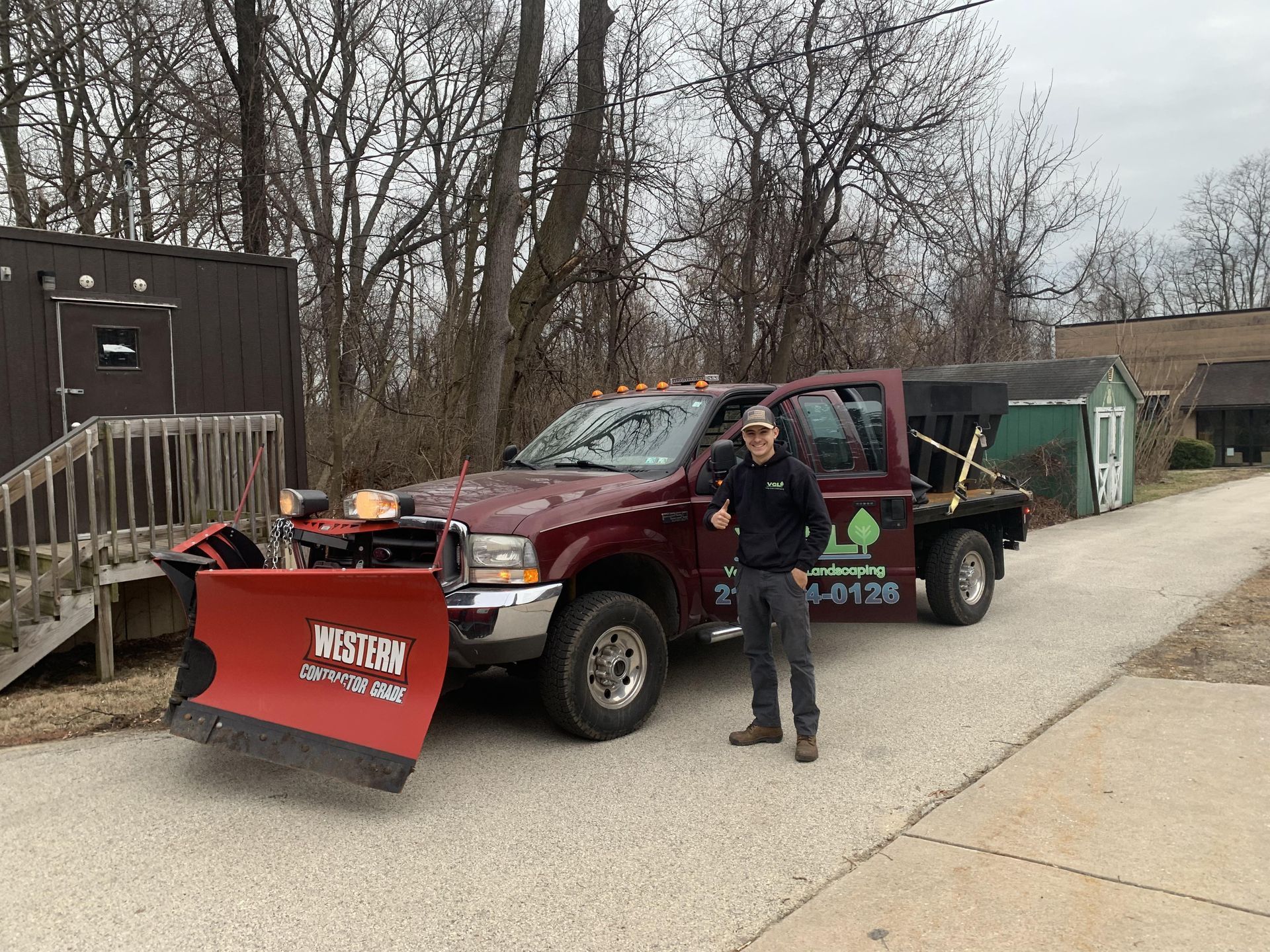 Man standing next to a burgundy snowplow truck on a paved road. Trees and a small building are in the background.