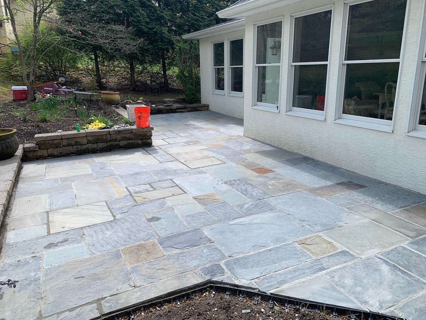Flagstone patio next to a white building with windows, bordering a small garden and trees.