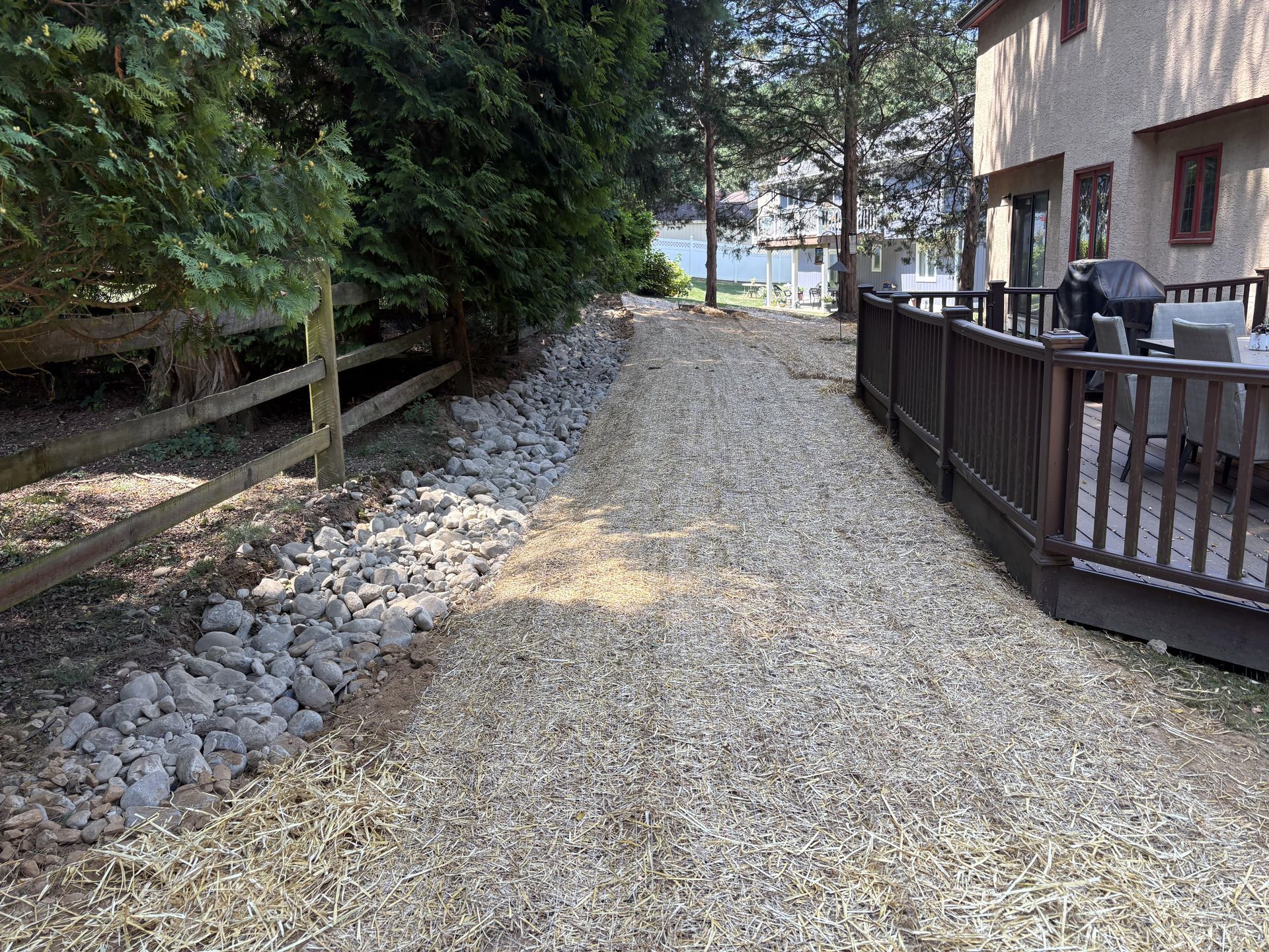 Gravel pathway alongside a wooden fence and a house with a deck, trees and water in the background.