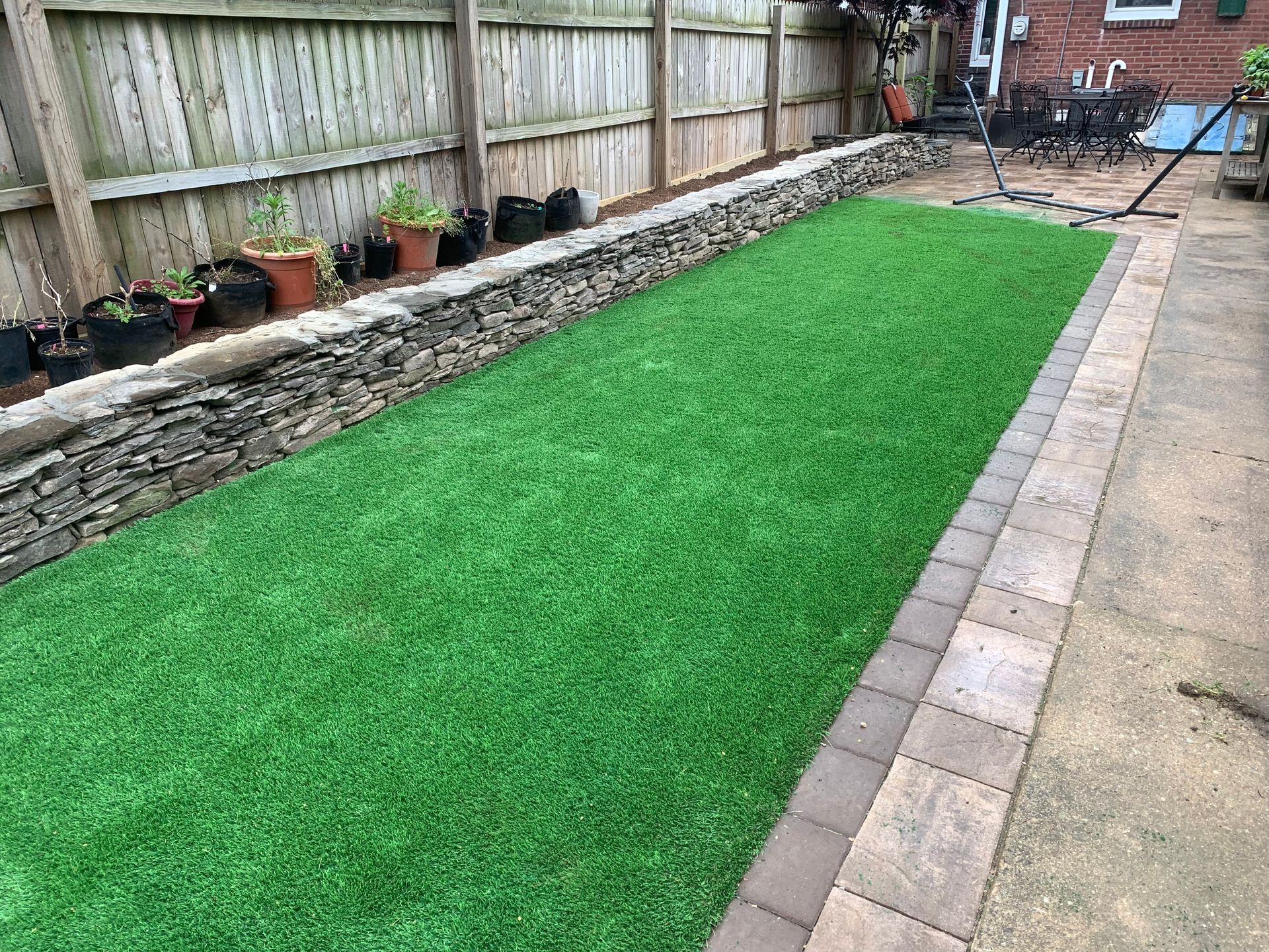 Long, narrow yard with green turf, bordered by stone wall and pavers, with wooden fence.