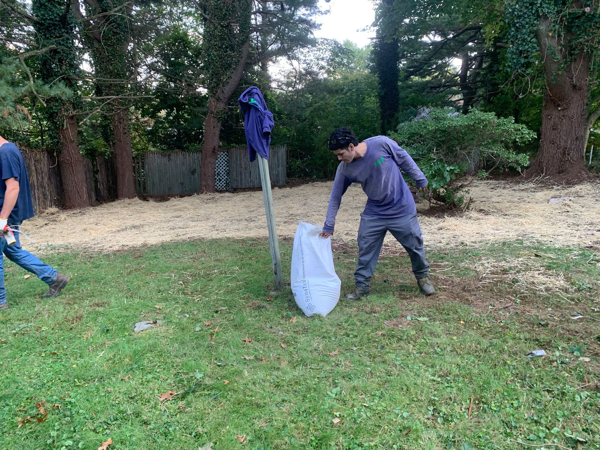 Person picking up a bag in a grassy yard, another person walks in background. Clothes hang on a post.