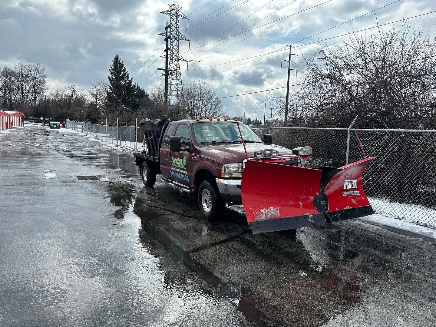 Red snowplow truck clearing snow from a wet asphalt lot near a chain-link fence and power lines.