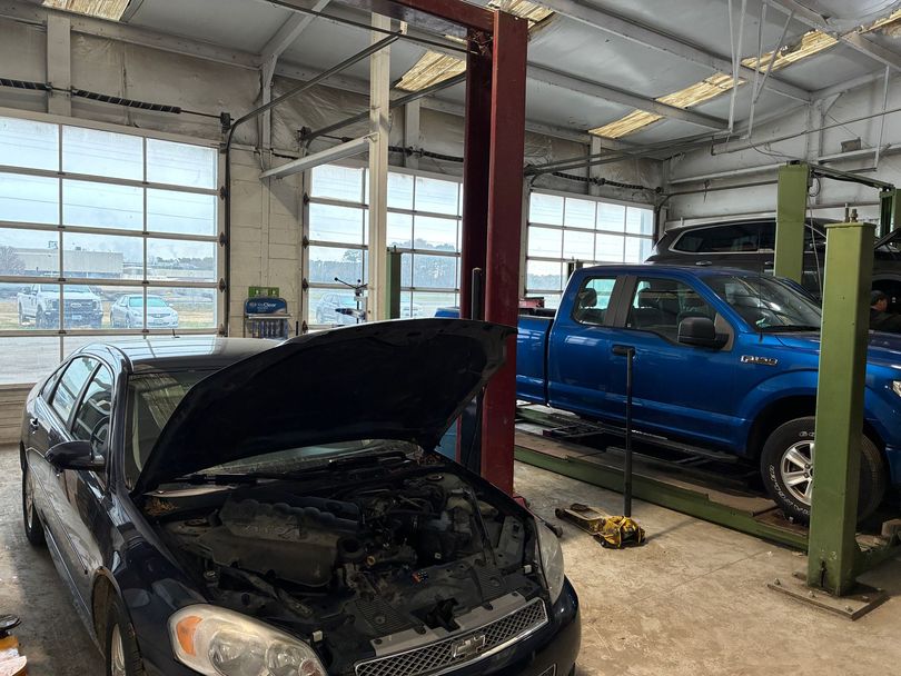 An auto repair shop with a dark blue car and a blue pickup truck on a lift. The car hood is open.