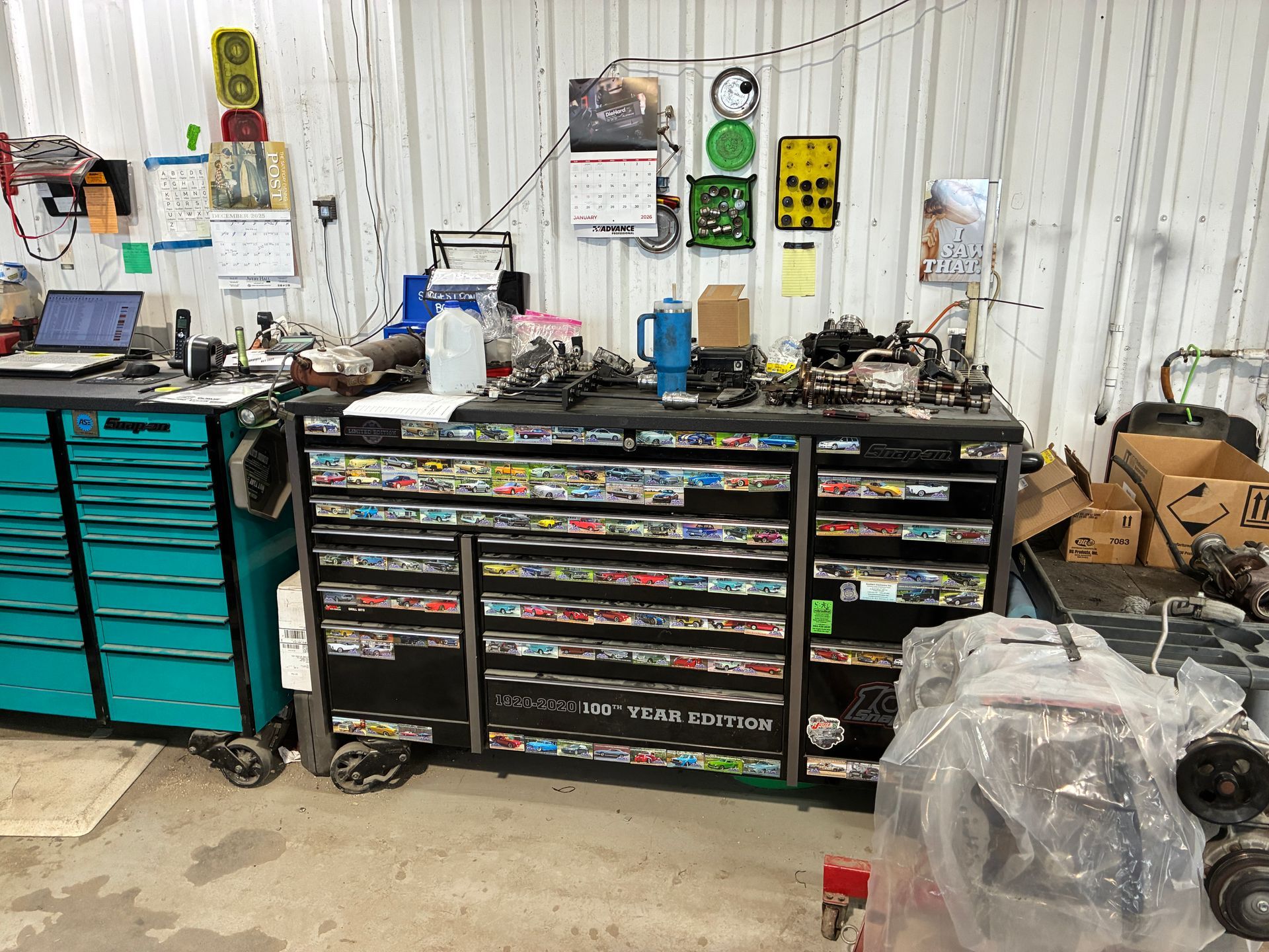 A cluttered garage workspace with a large black tool chest, teal cabinet, and engine parts.