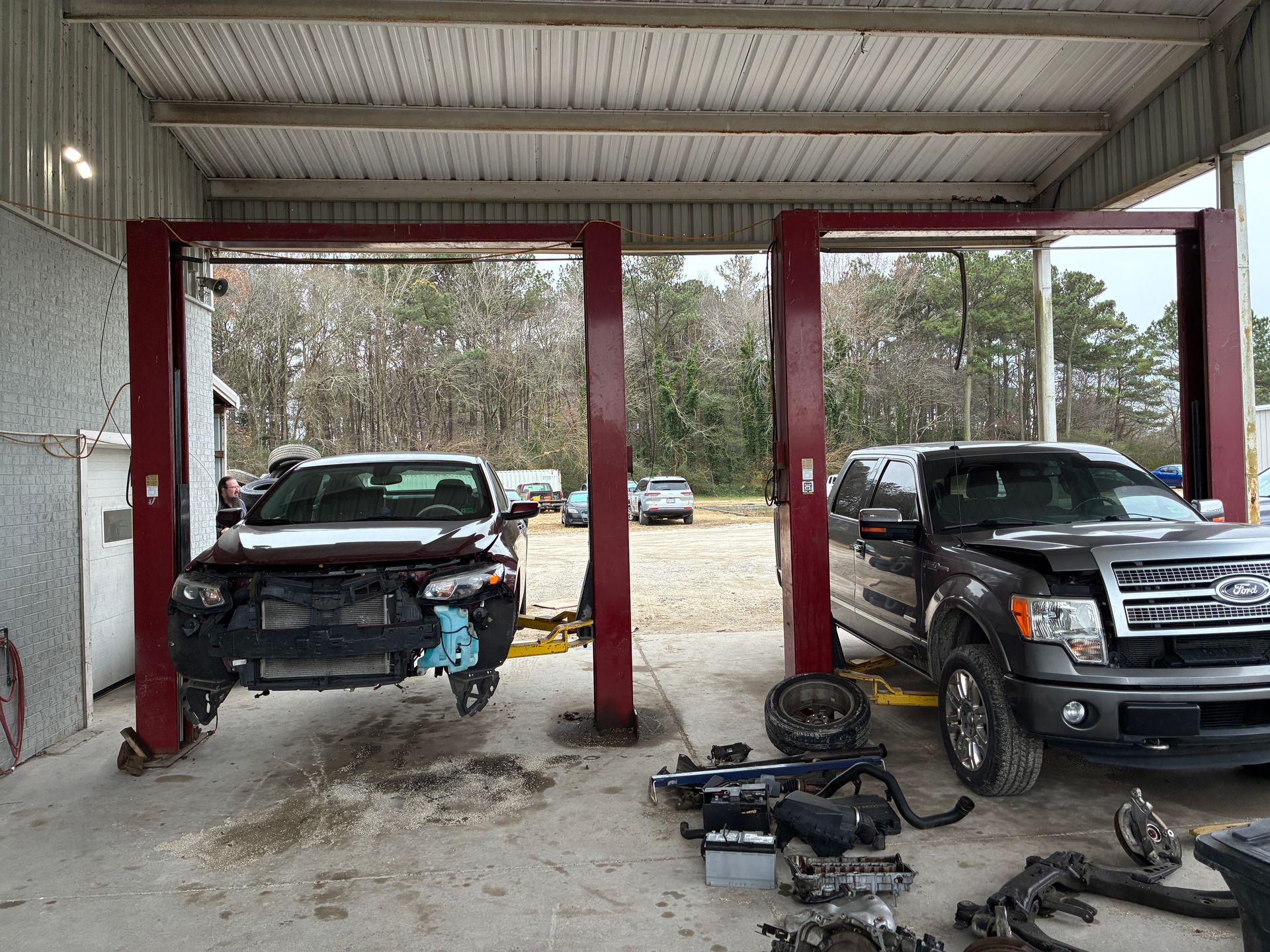 Two vehicles being repaired in an auto shop. One is raised on a lift with front end damage. Tools and parts are scattered on the floor.