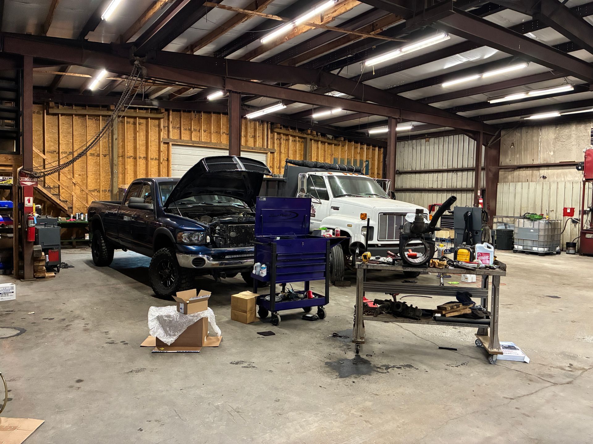 A dark blue pickup truck with its hood open and a white truck parked inside a repair shop.