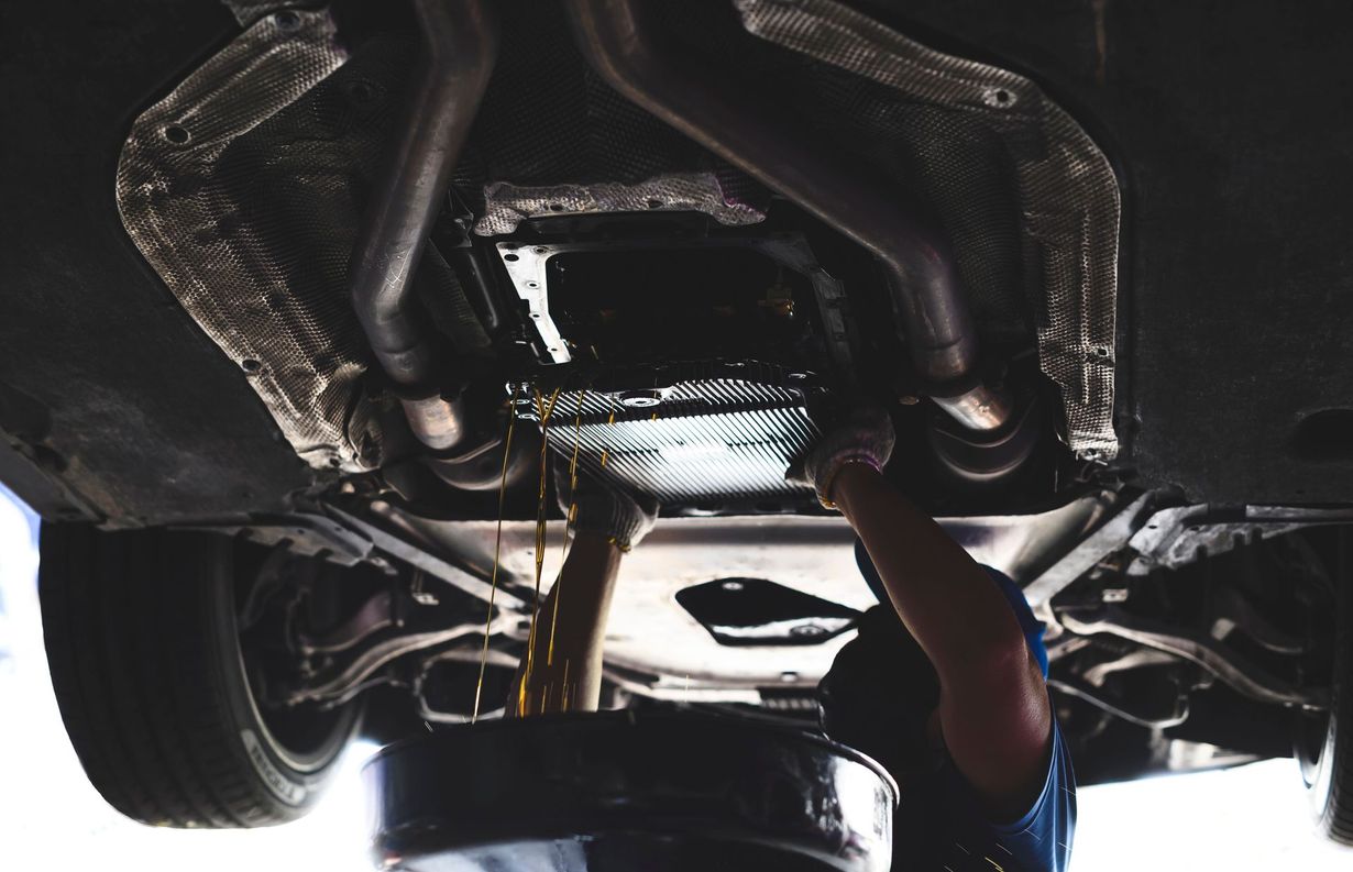 Mechanic draining oil from a car. Oil pan under vehicle, exhaust pipes, setting is a garage.