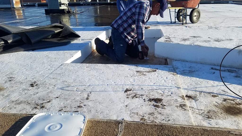 A man is working on a roof with a wheelbarrow in the background.