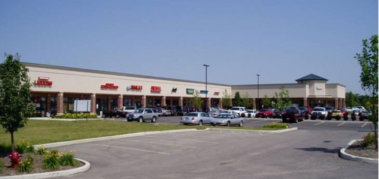 Shopping plaza with storefronts, parking lot, and parked cars under a blue sky.