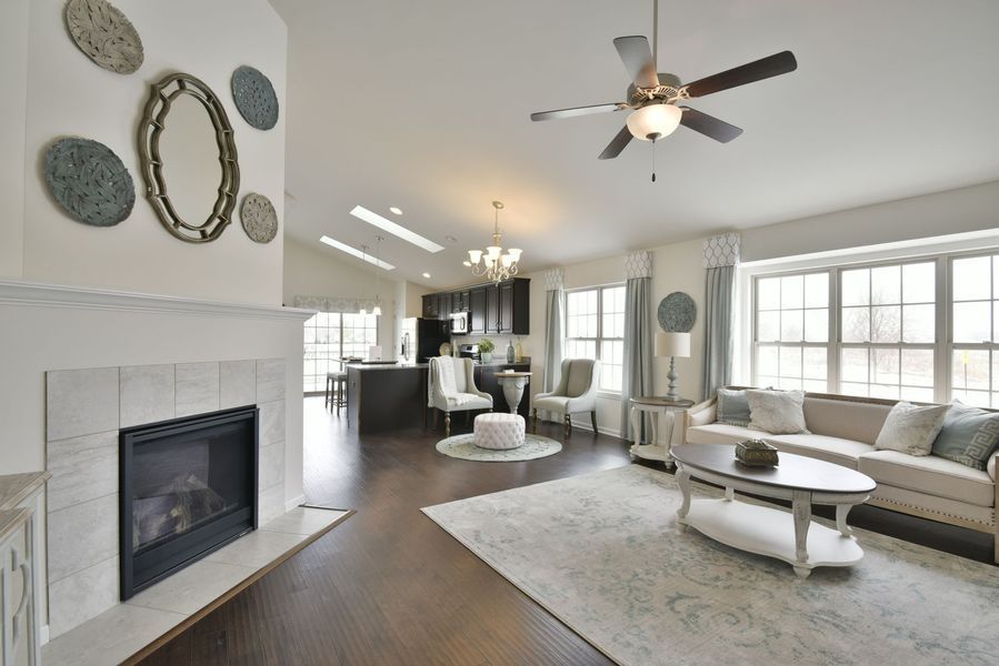 Living room with fireplace, sofa, and kitchen in the background. Light-filled with dark wood floors and white walls.