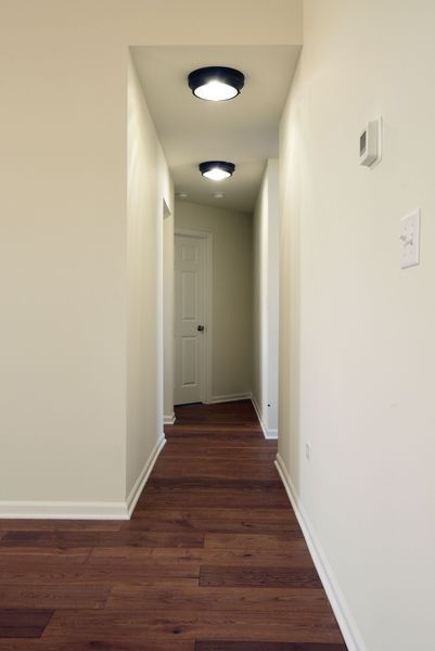 Narrow hallway with dark wood flooring, white walls, and overhead lighting. A closed white door is at the end.