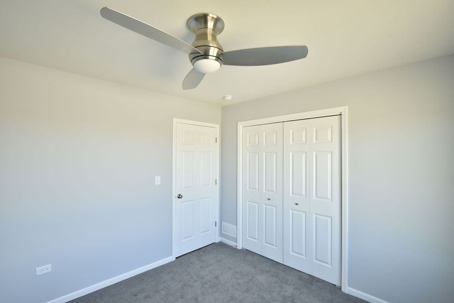 Empty bedroom with a ceiling fan, closet, and a door. Light gray walls and carpet.