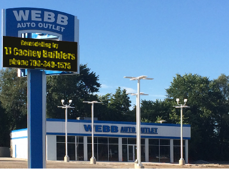 Webb Auto Outlet sign and building against a blue sky; display lists the builders' name and phone number.
