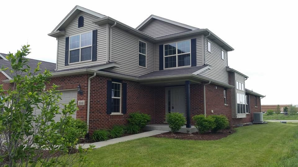 Two-story house with brick exterior, tan siding, and dark gray roof. Green lawn and overcast sky.