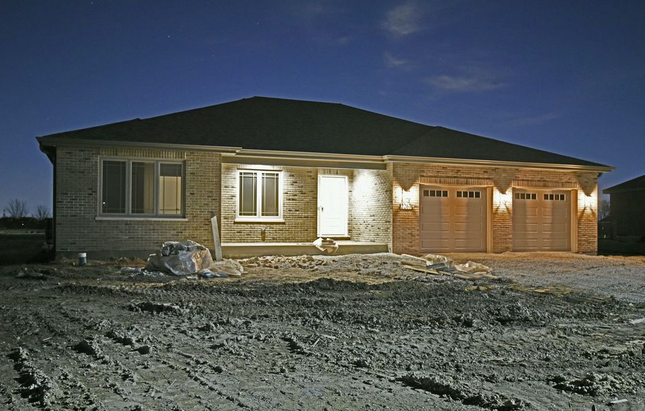 House at night with exterior lights on. Tan brick facade, dark roof, two garage doors, and unfinished yard.