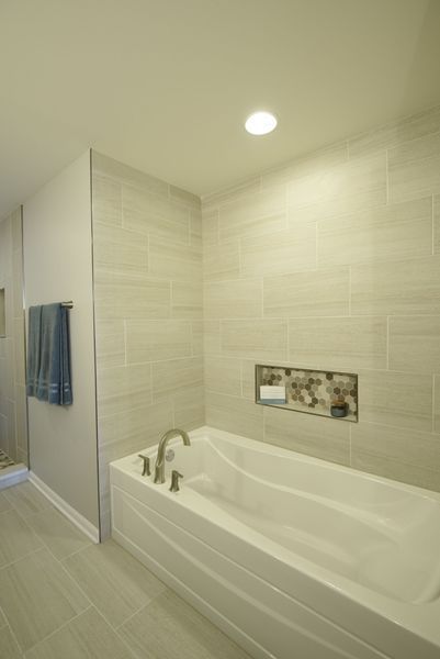 Bathroom with a white bathtub, light gray tile walls, and a recessed shelf. Blue towels hang on the wall.