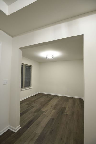 Empty room with hardwood floors, a window, and overhead lighting, viewed through an open doorway.