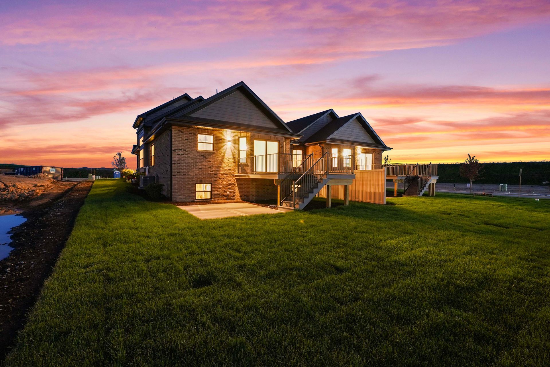 Brick house with warm lights at sunset, beside a grassy yard under a pink sky.