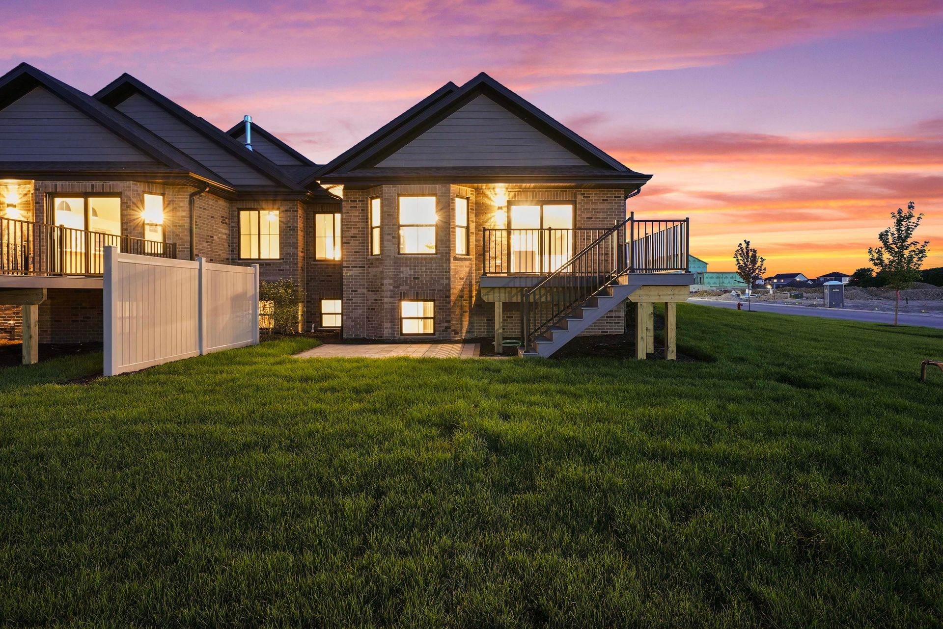 Modern house with lit windows at sunset, overlooking a grassy yard under a pink-orange sky