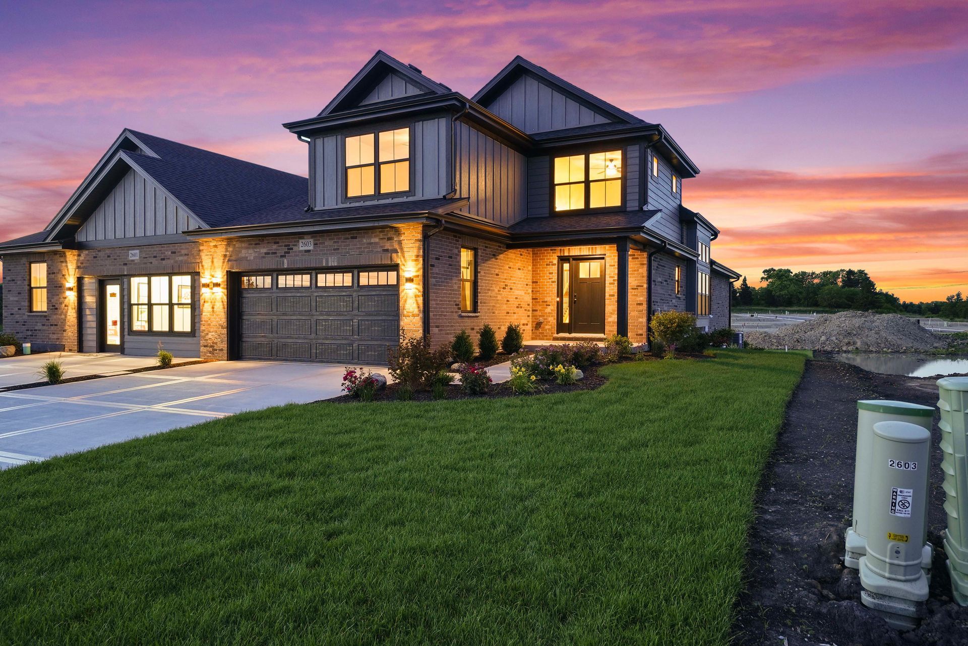 Large modern house at dusk with lit windows, a driveway, and a green lawn under a pink-purple sky