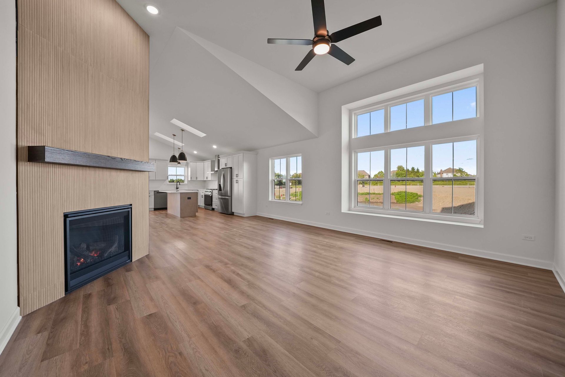 Bright empty living room with wood floors, fireplace, high ceiling fan, and large windows