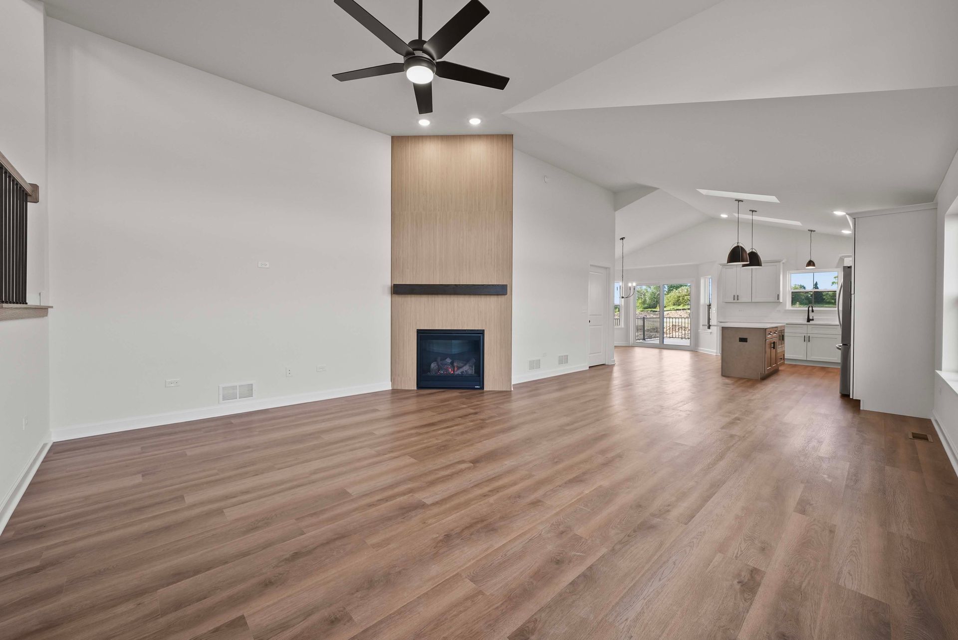 Spacious empty living room with wood floors, white walls, and a fireplace opening to a bright kitchen.