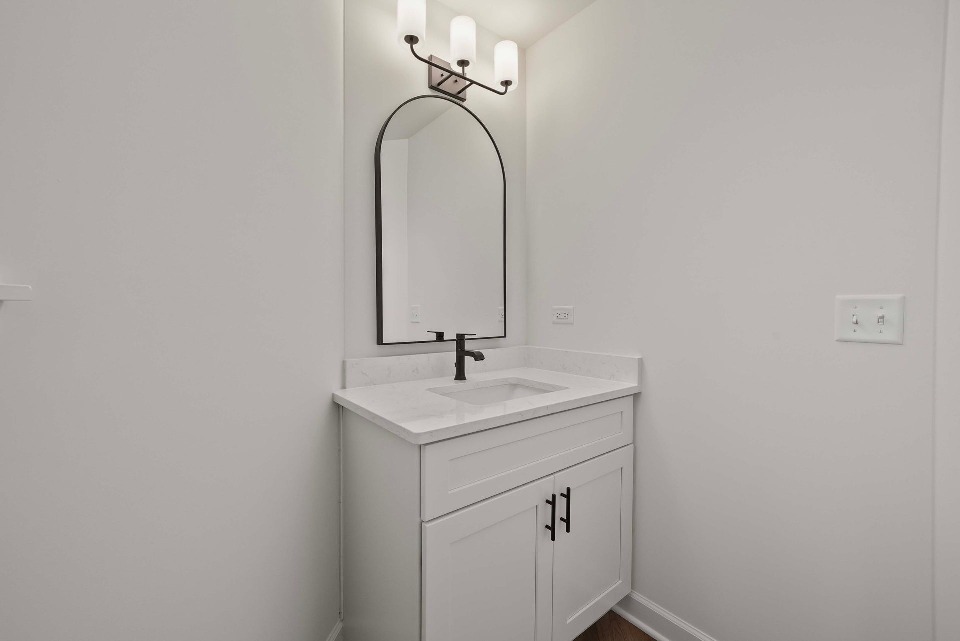 Small white bathroom vanity with sink, arched mirror, and wall sconce in a bright corner.