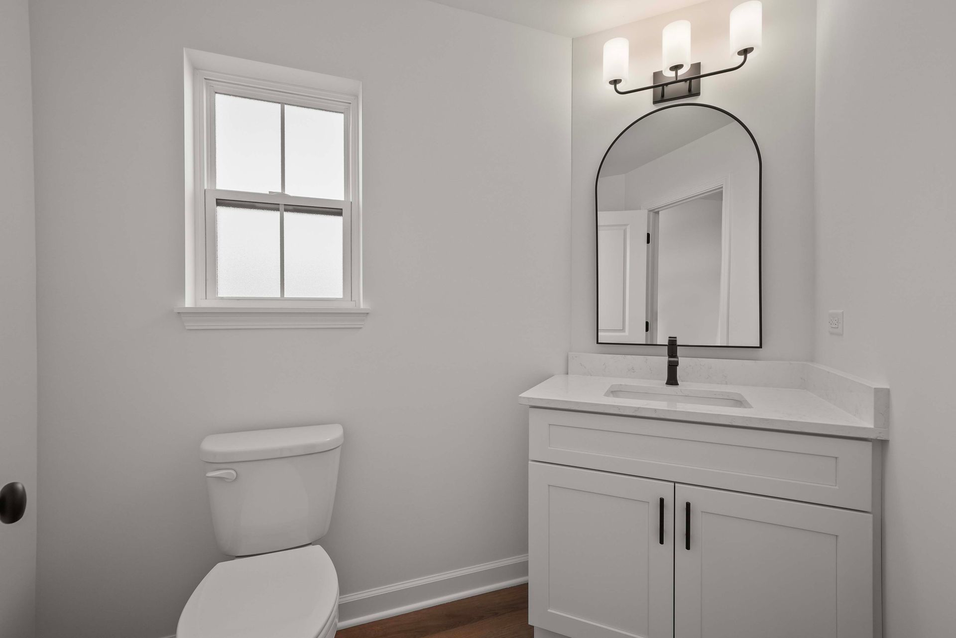 Small white bathroom with a toilet, window, vanity sink, and arched mirror.