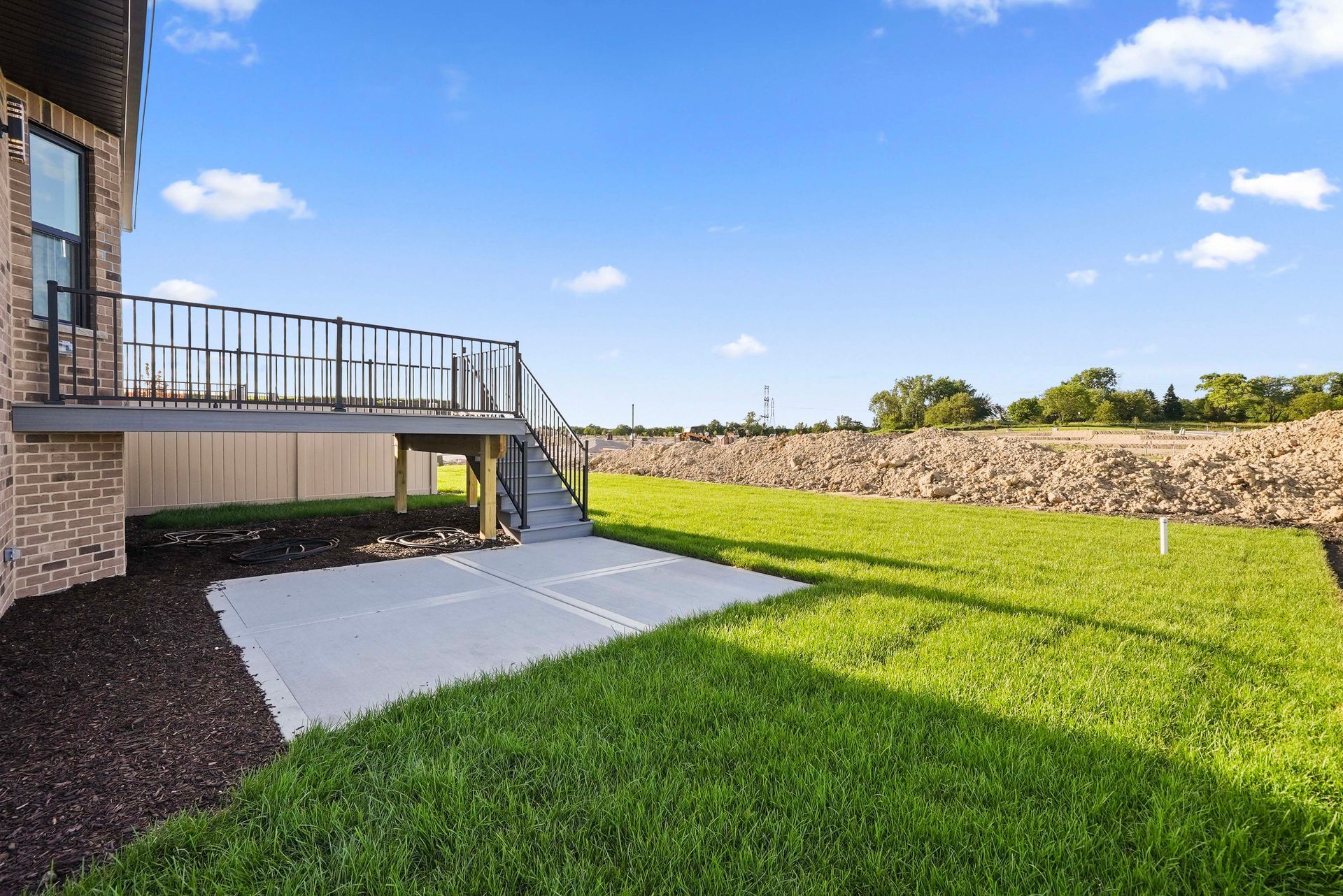 Backyard patio with a raised deck, green lawn, and stone retaining wall under a blue sky