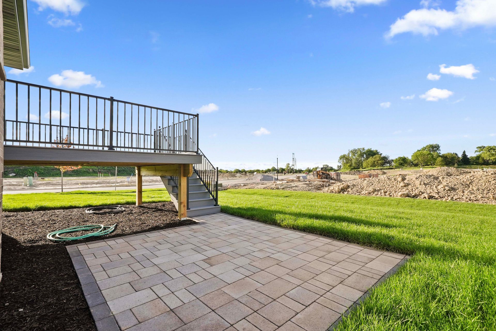 Backyard patio with stone pavers, grassy lawn, and raised wooden deck under a blue sky
