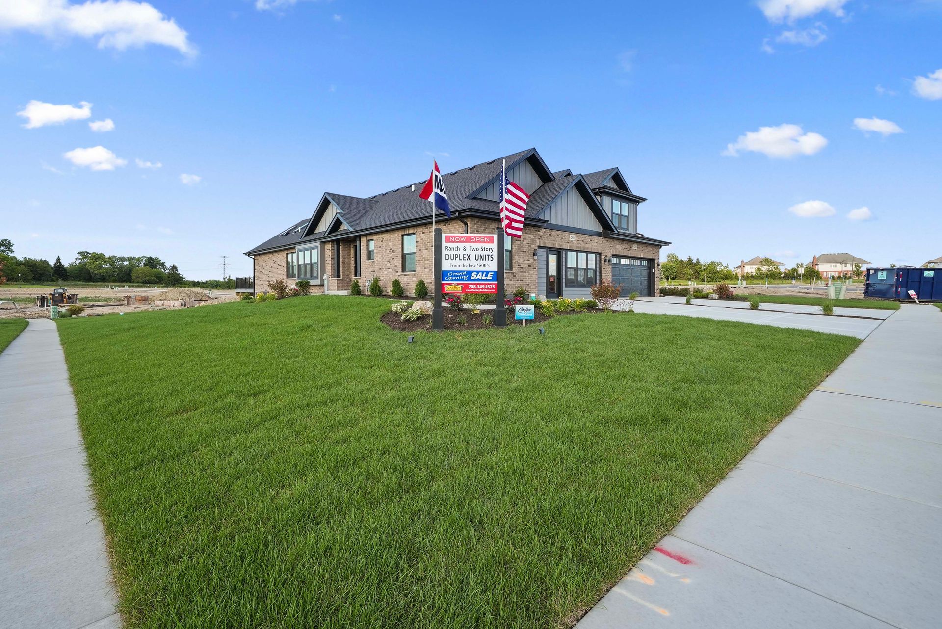 Stone house with American flags and a real estate sign on a grassy corner lot under a blue sky