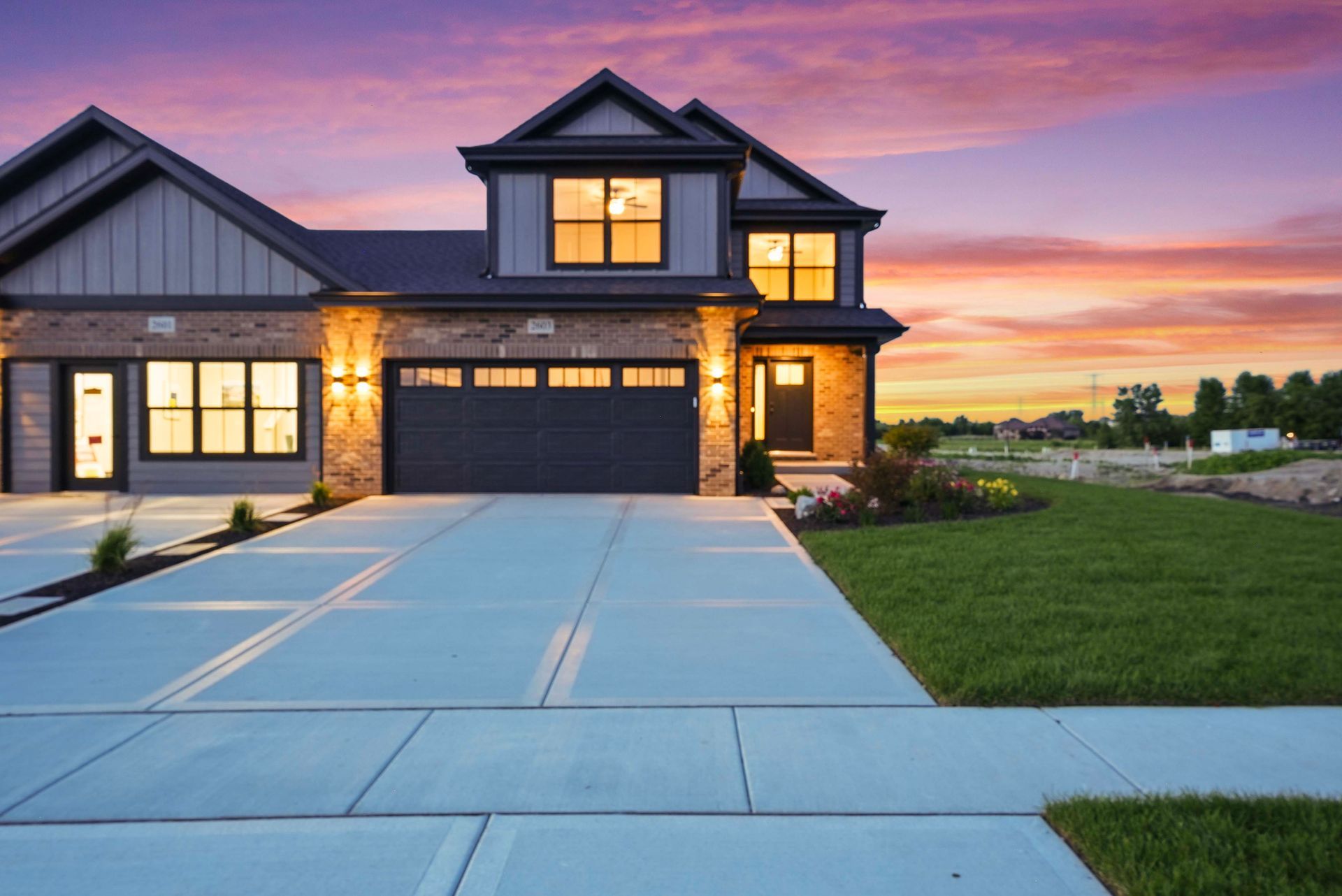 Modern two-story house with lit windows, stone accents, and a wide driveway at sunset