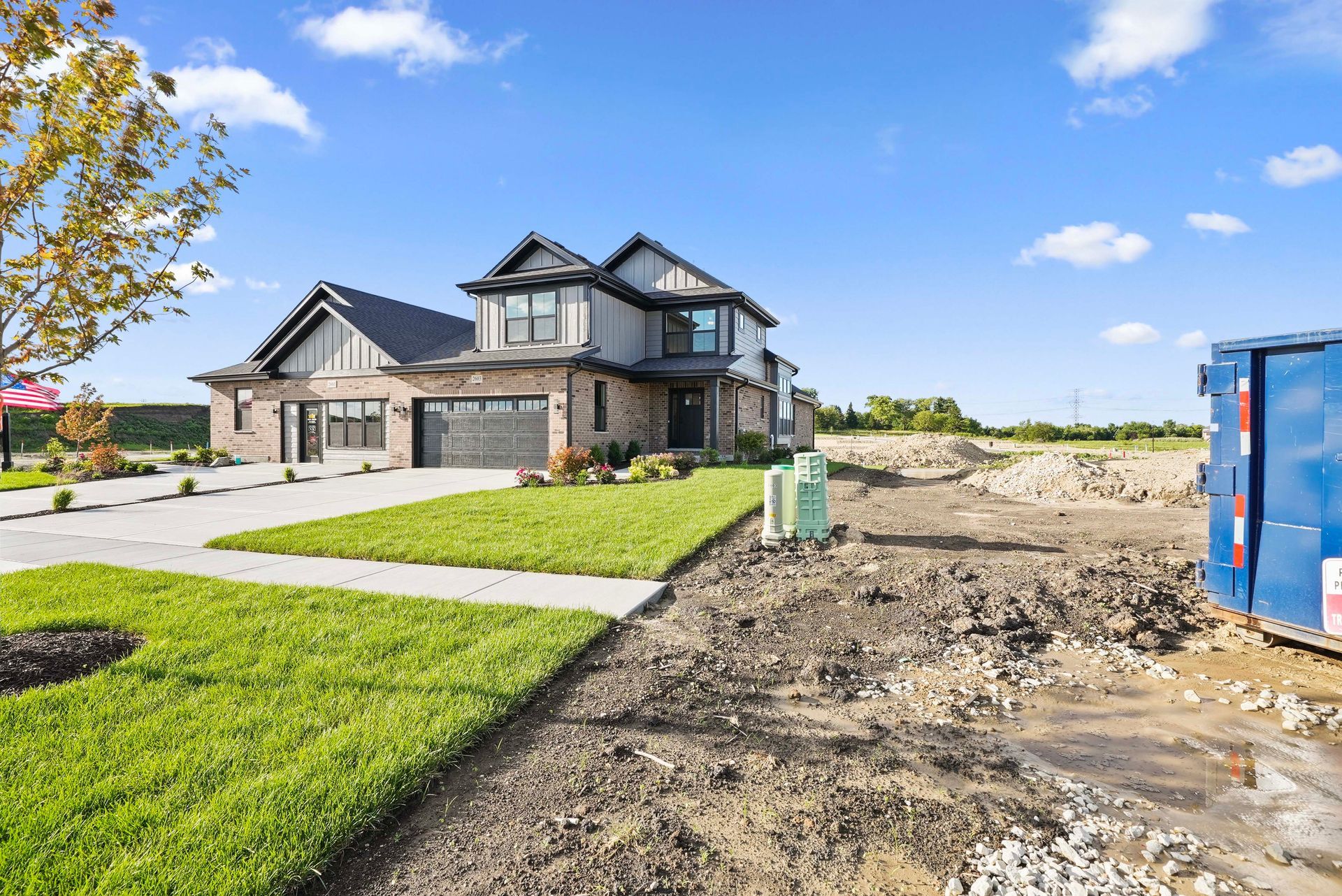 New house under construction beside a paved driveway and dirt lot, with a blue dumpster on the right