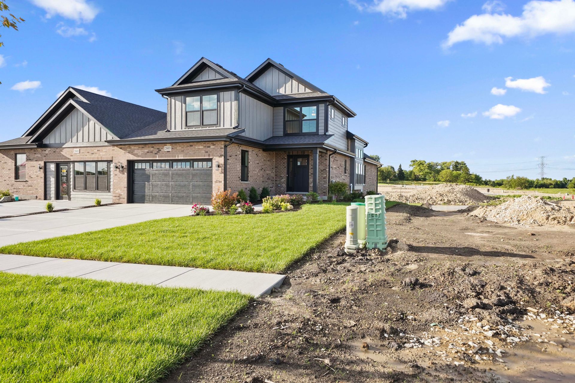 New brick house with black roof, green lawn, and unfinished dirt lot under a blue sky