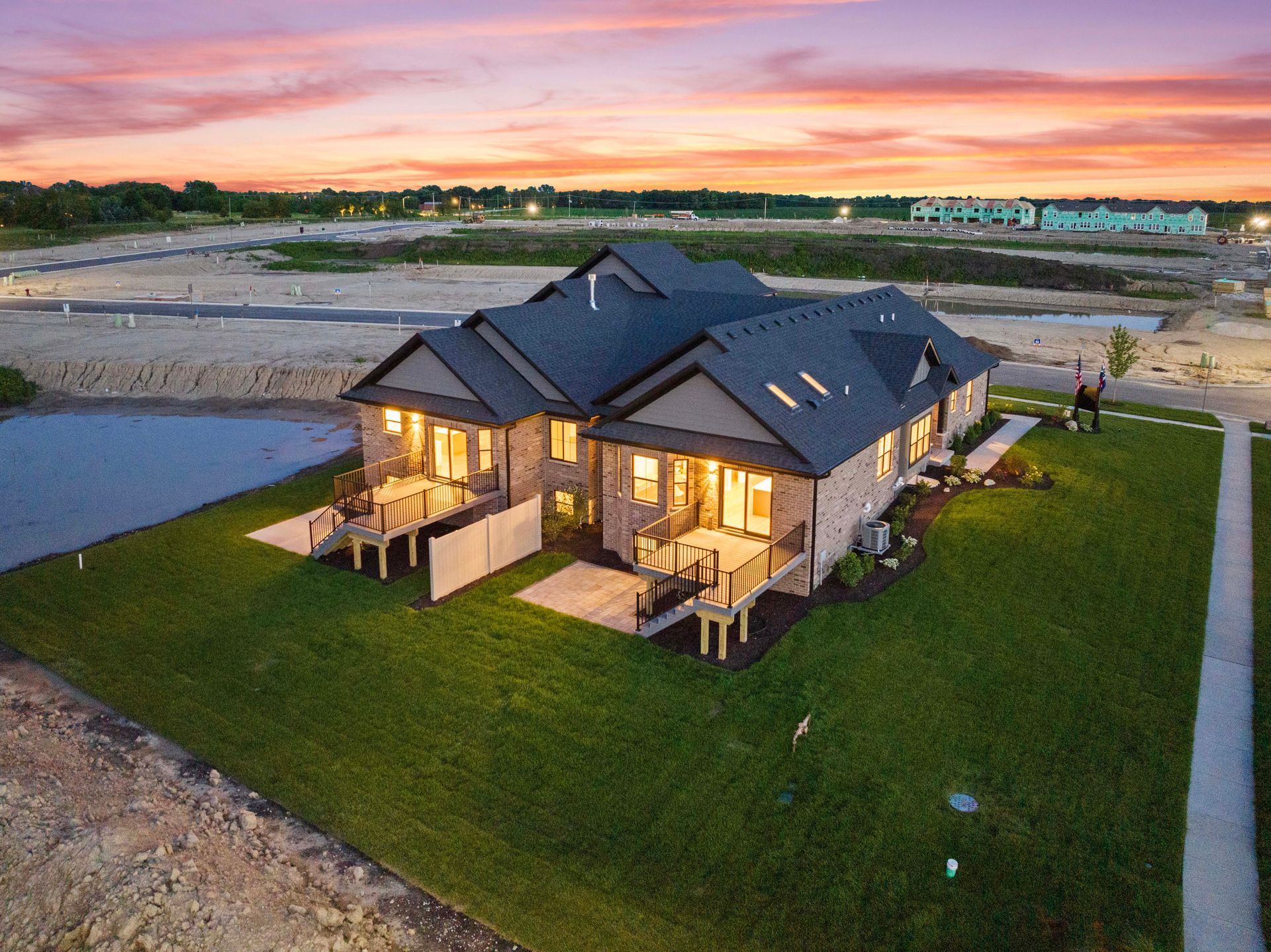 Aerial view of a modern house with warm interior lights, set on a green lot near water at sunset