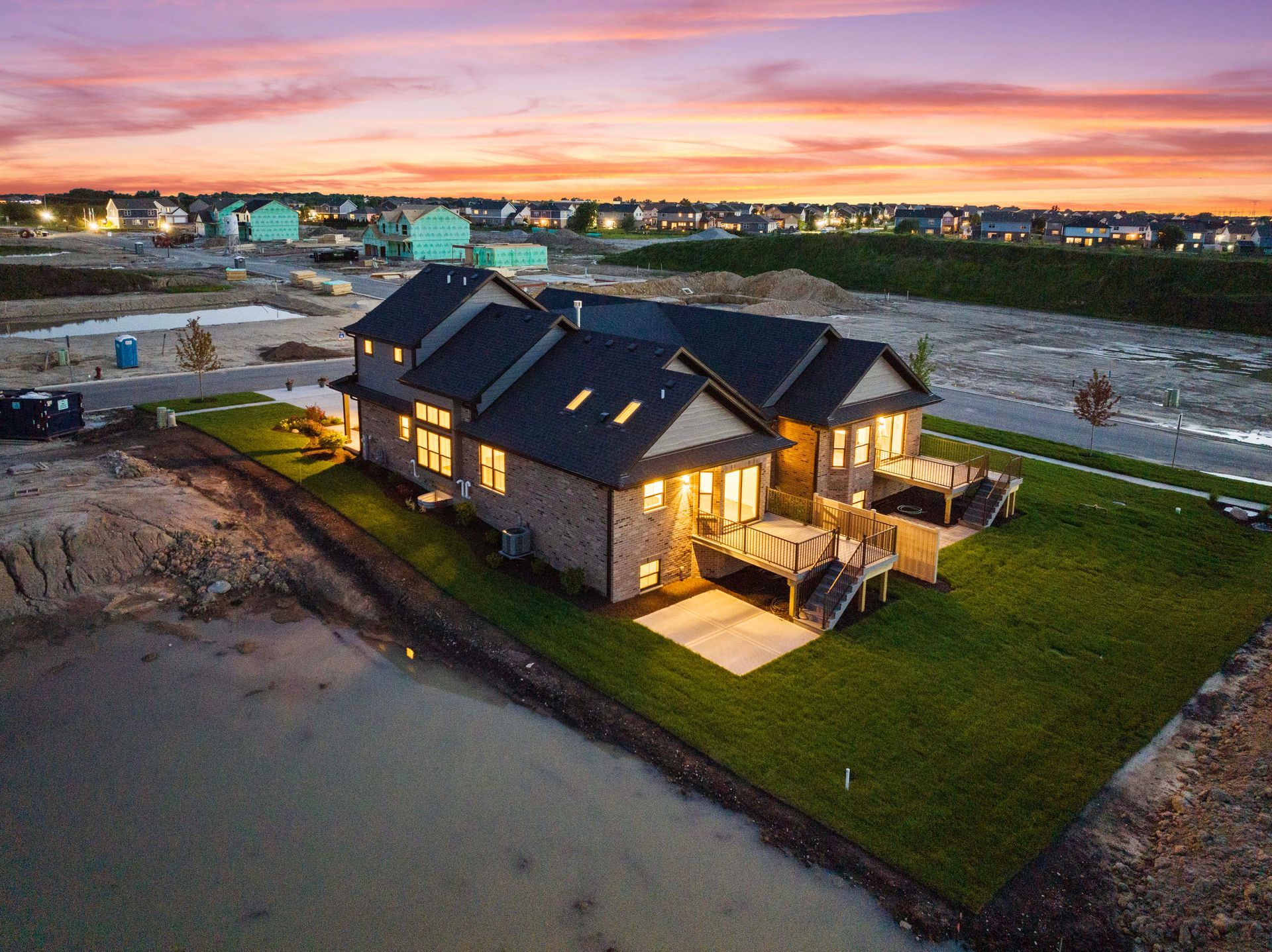 Modern house at dusk by a sandy shoreline, with warm lights and a glowing sunset sky