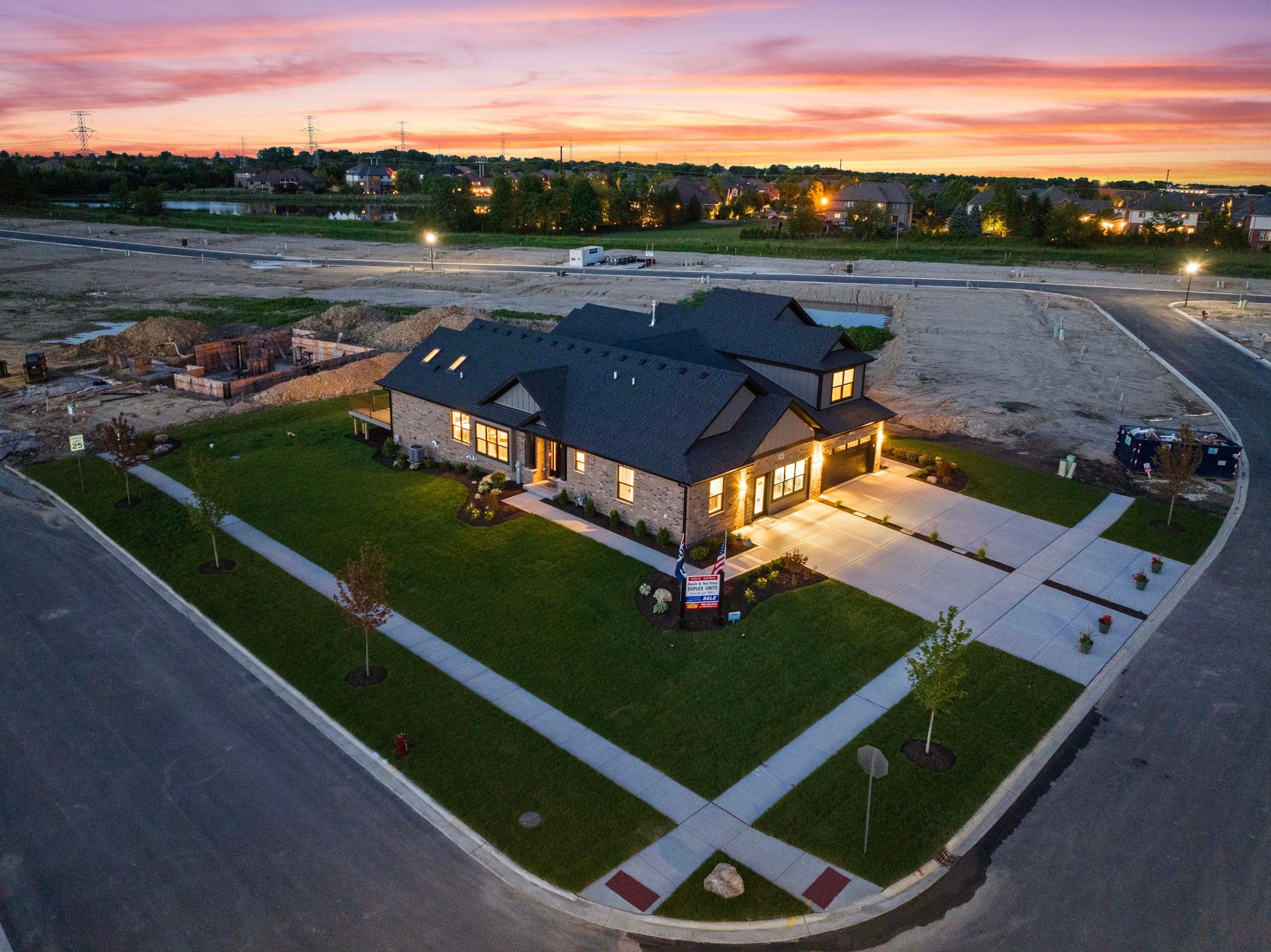 Aerial view of a lit modern house at dusk with a landscaped corner lot and sunset sky
