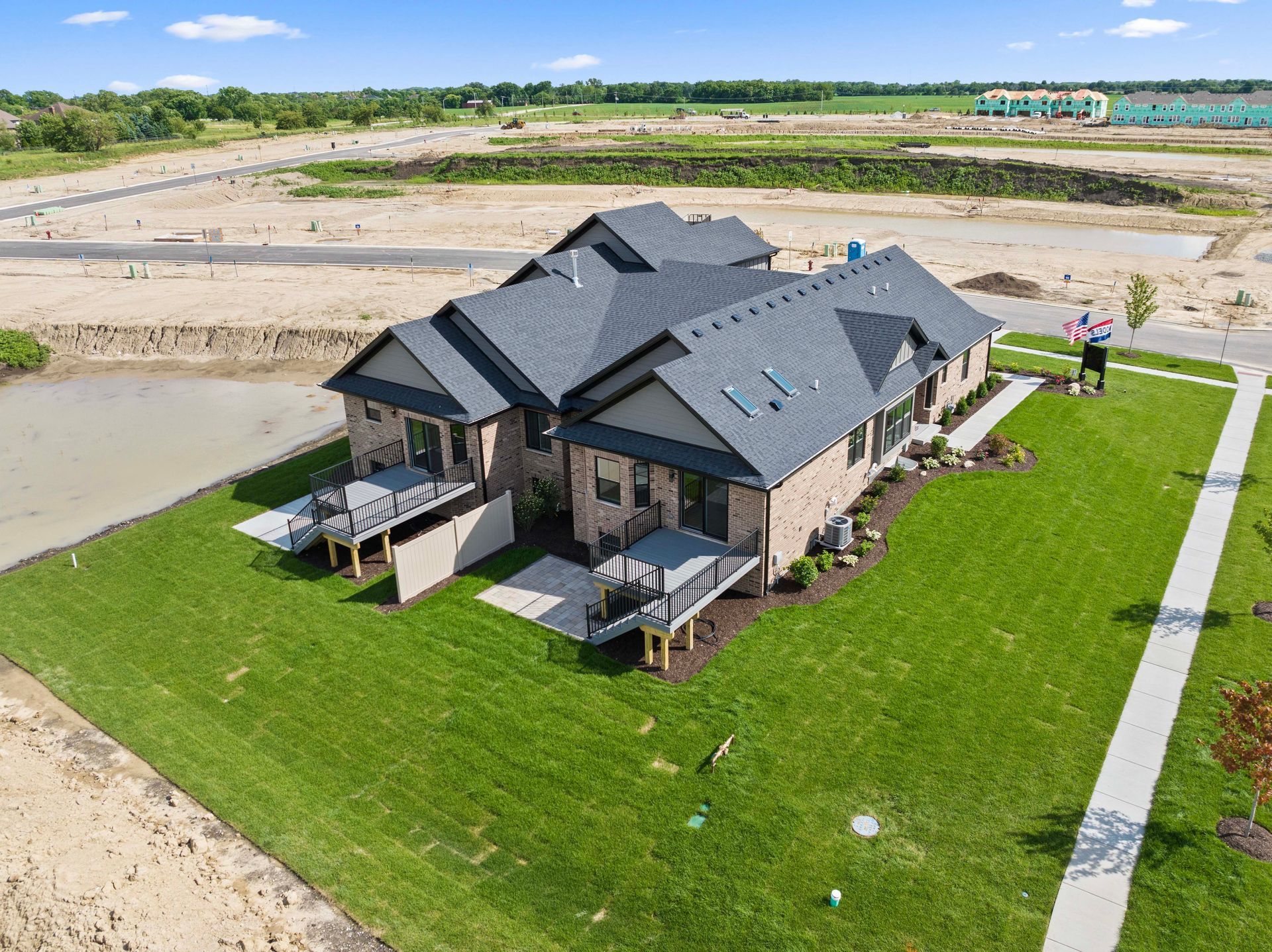 Aerial view of a large stone house with a dark roof, green lawn, and a nearby pond in a suburban development