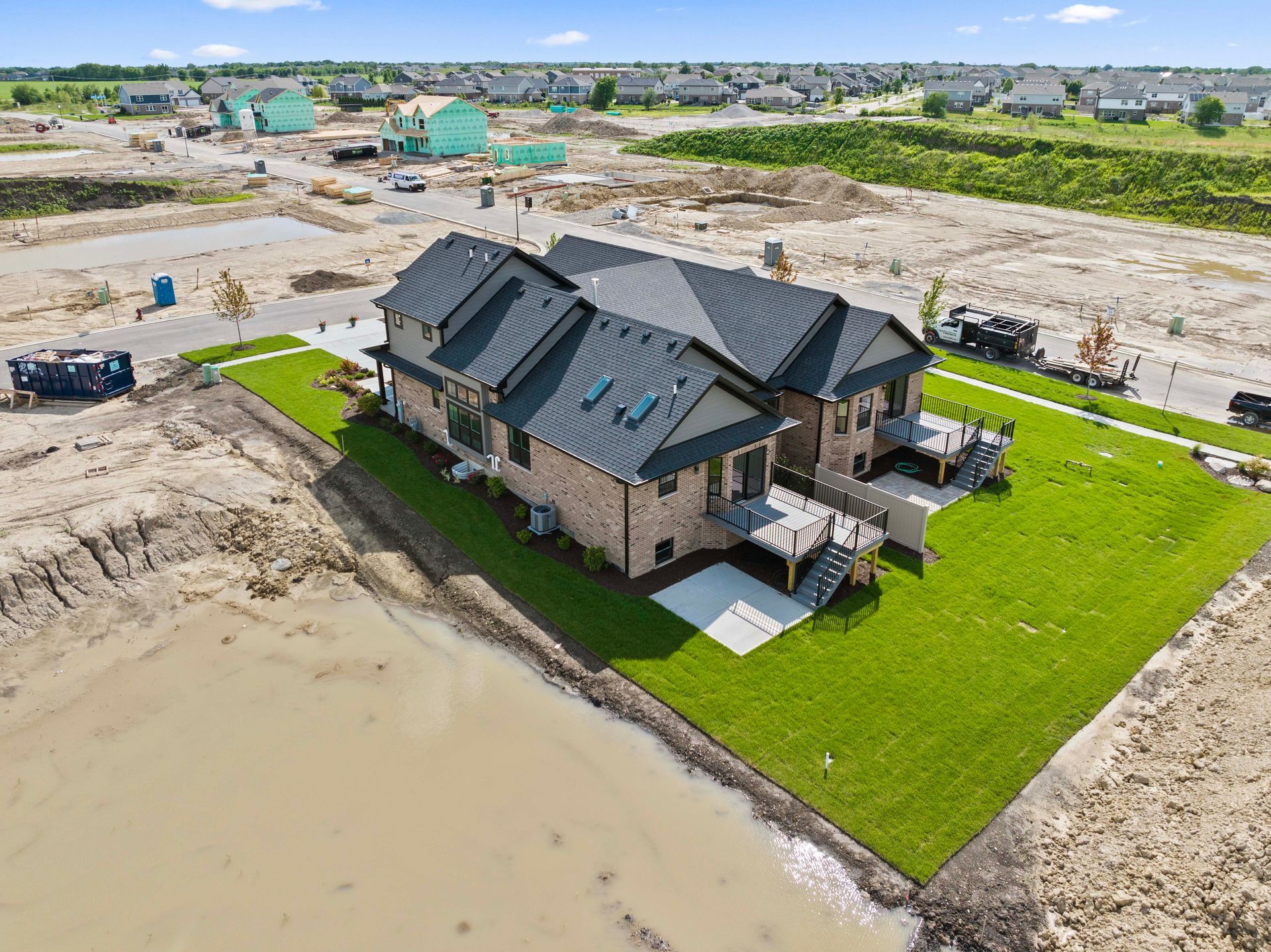 Aerial view of a large house beside a green lawn and muddy floodwater, with a construction site nearby