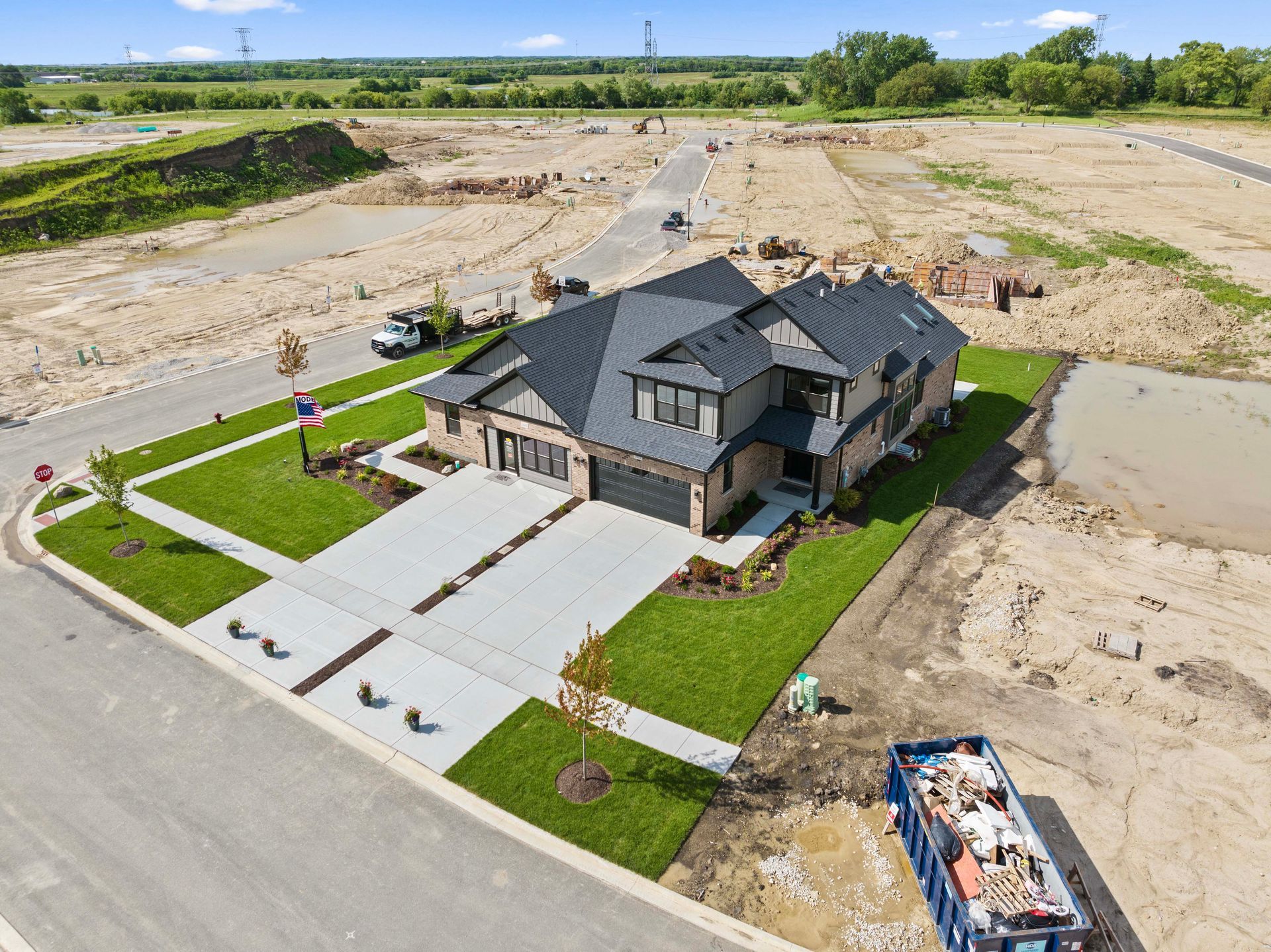 Aerial view of a new dark-gray house with a paved driveway, green lawn, and surrounding construction site