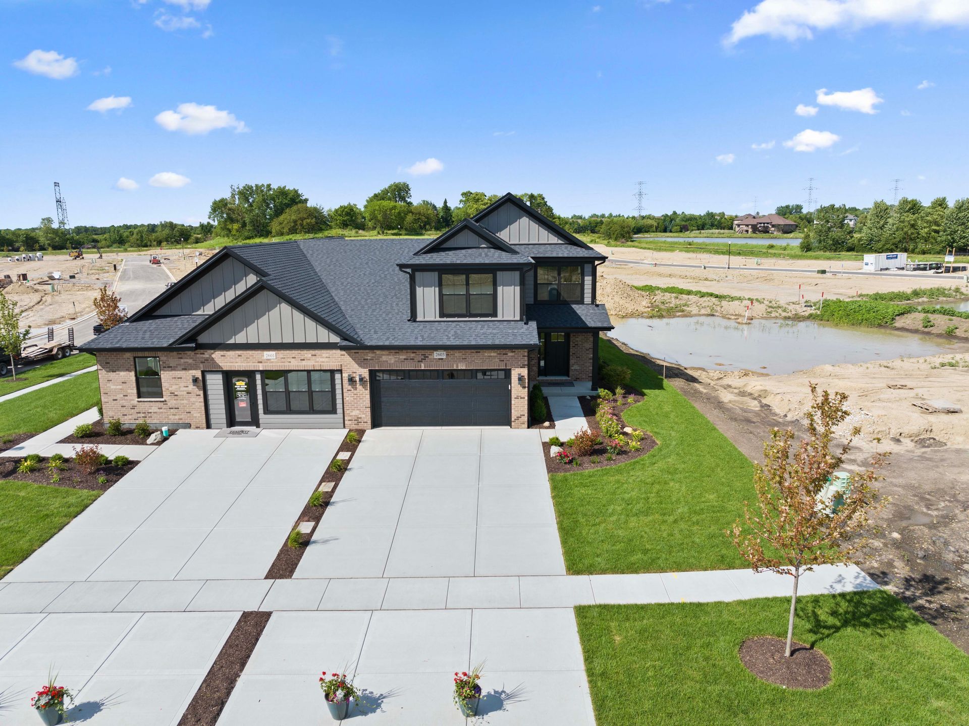 Modern two-story house with dark roof, large driveway, and lawn beside a barren construction lot under a blue sky