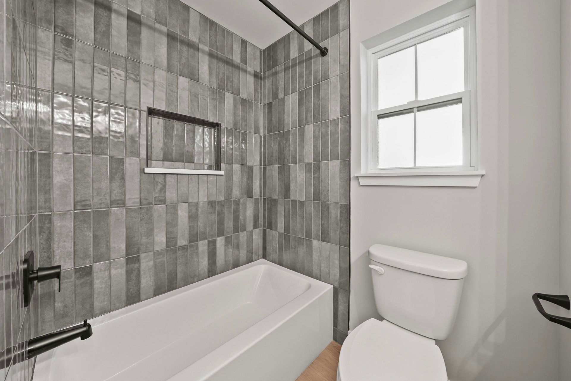 Small bathroom with gray tile shower, white tub, toilet, and window.