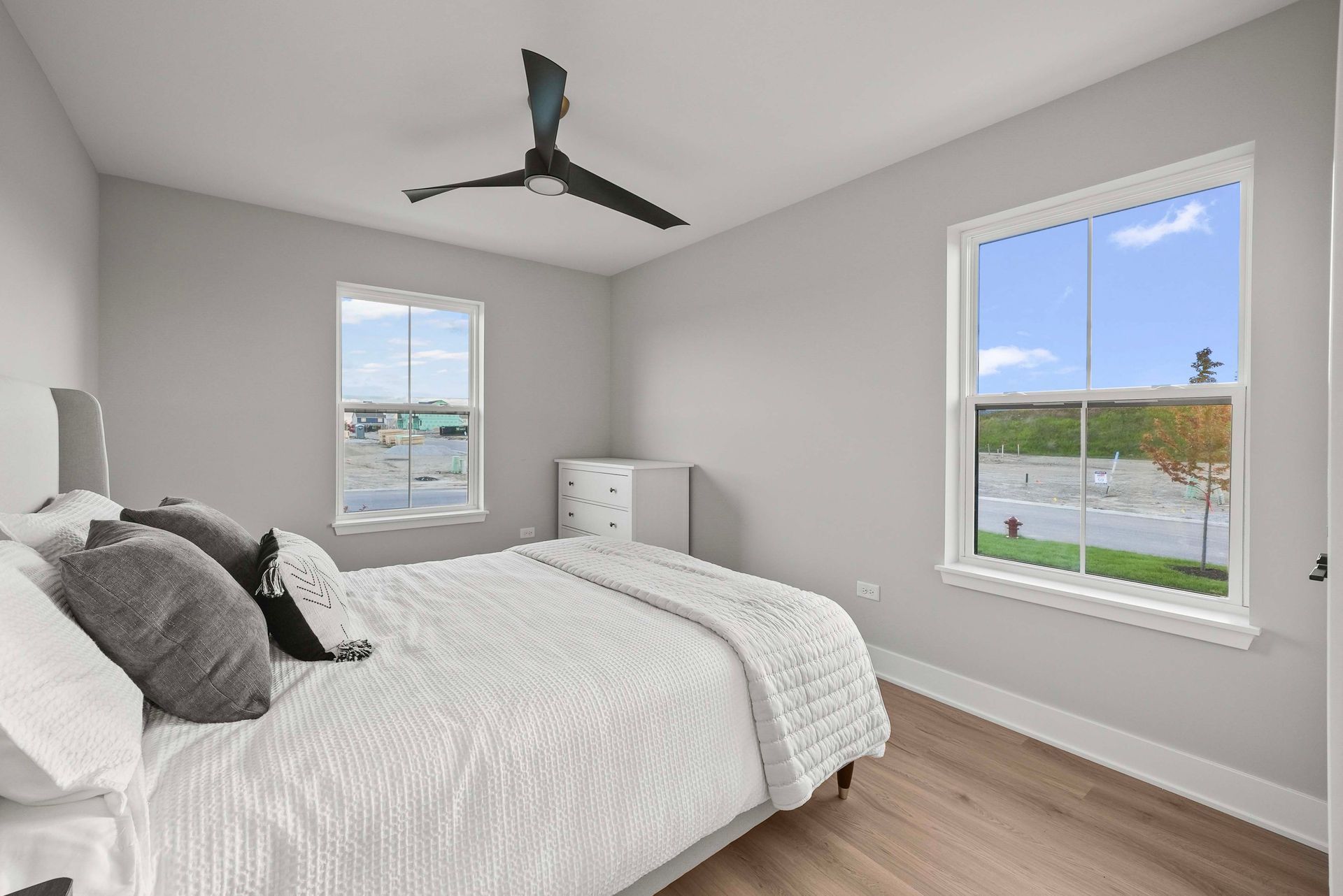Bright bedroom with white bedding, gray walls, wood floor, ceiling fan, and two windows.
