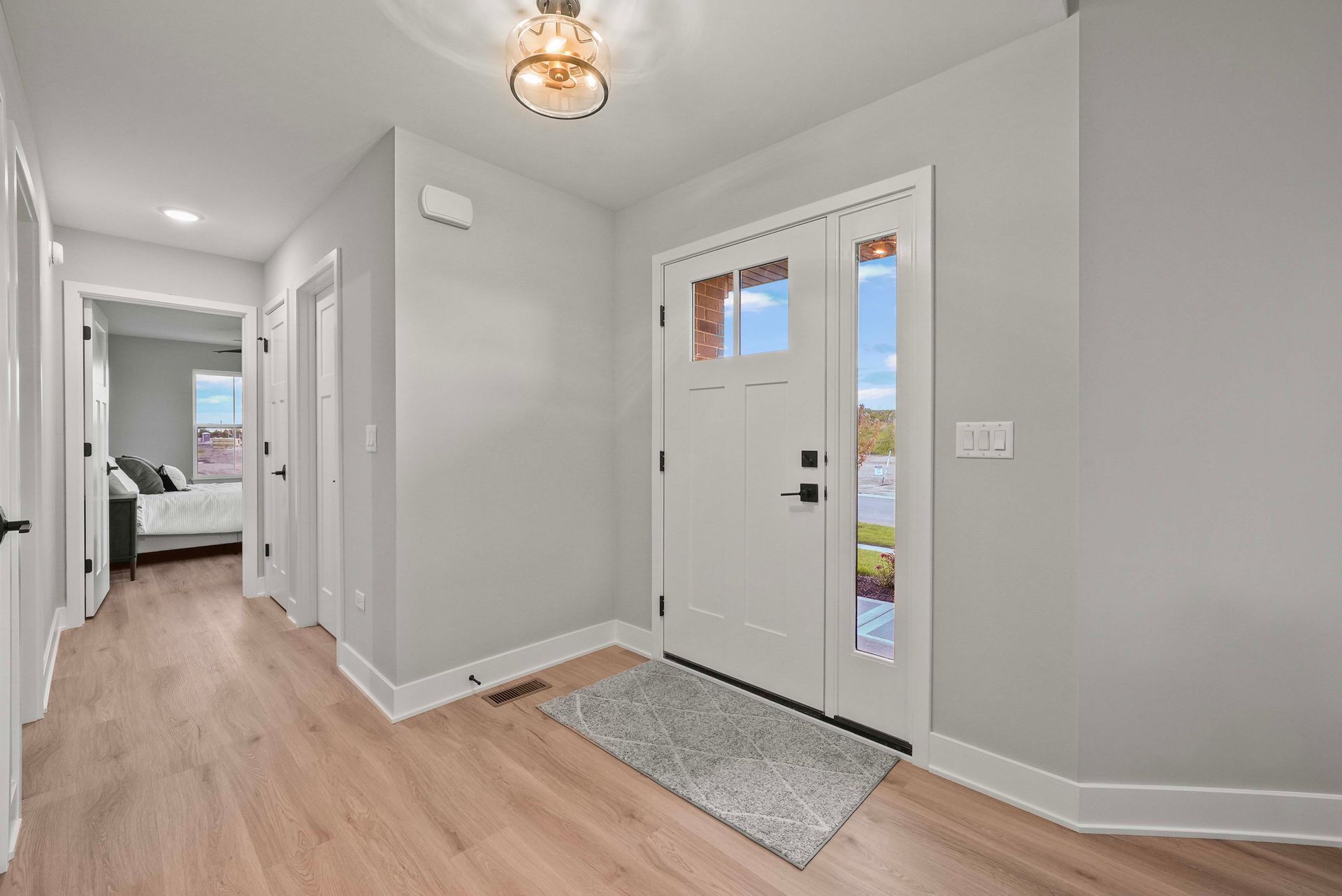 Bright entryway with white front door, gray walls, light wood floor, and a view into a bedroom hallway.