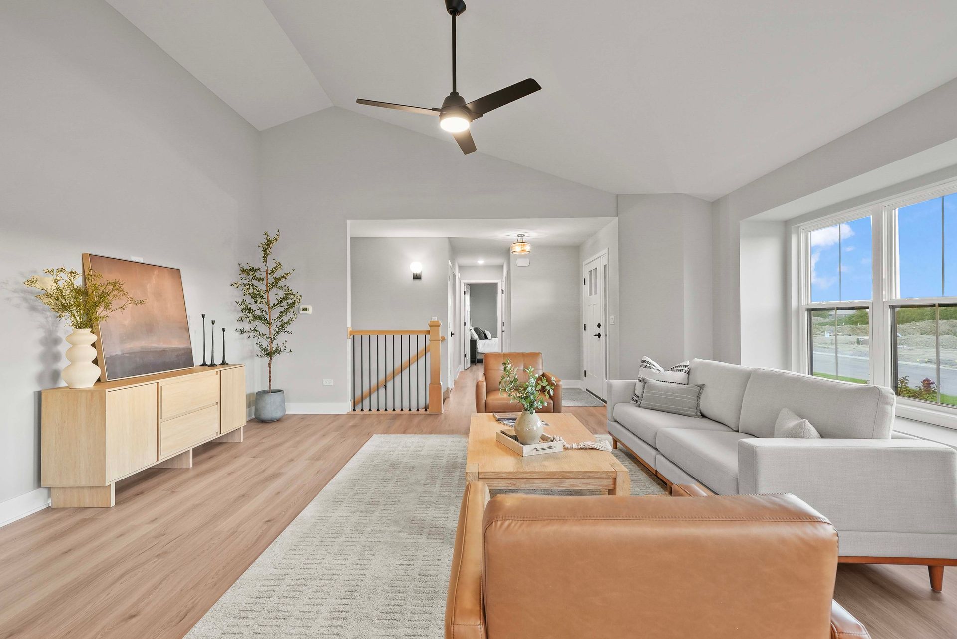 Bright living room with gray sofa, tan chair, wood floor, and large windows under a vaulted ceiling.