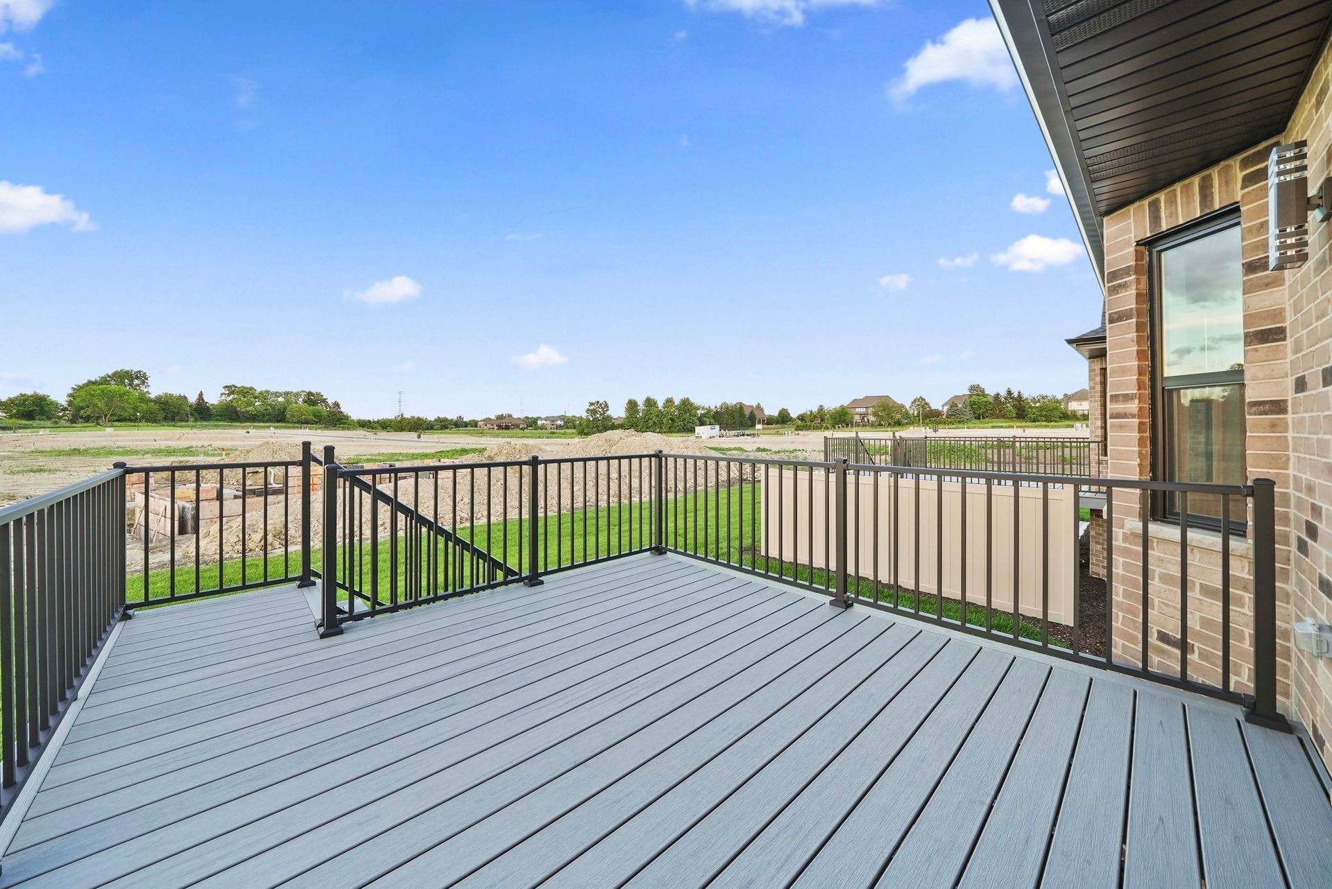 Spacious gray deck with black railings beside a brick house, overlooking open fields under a blue sky