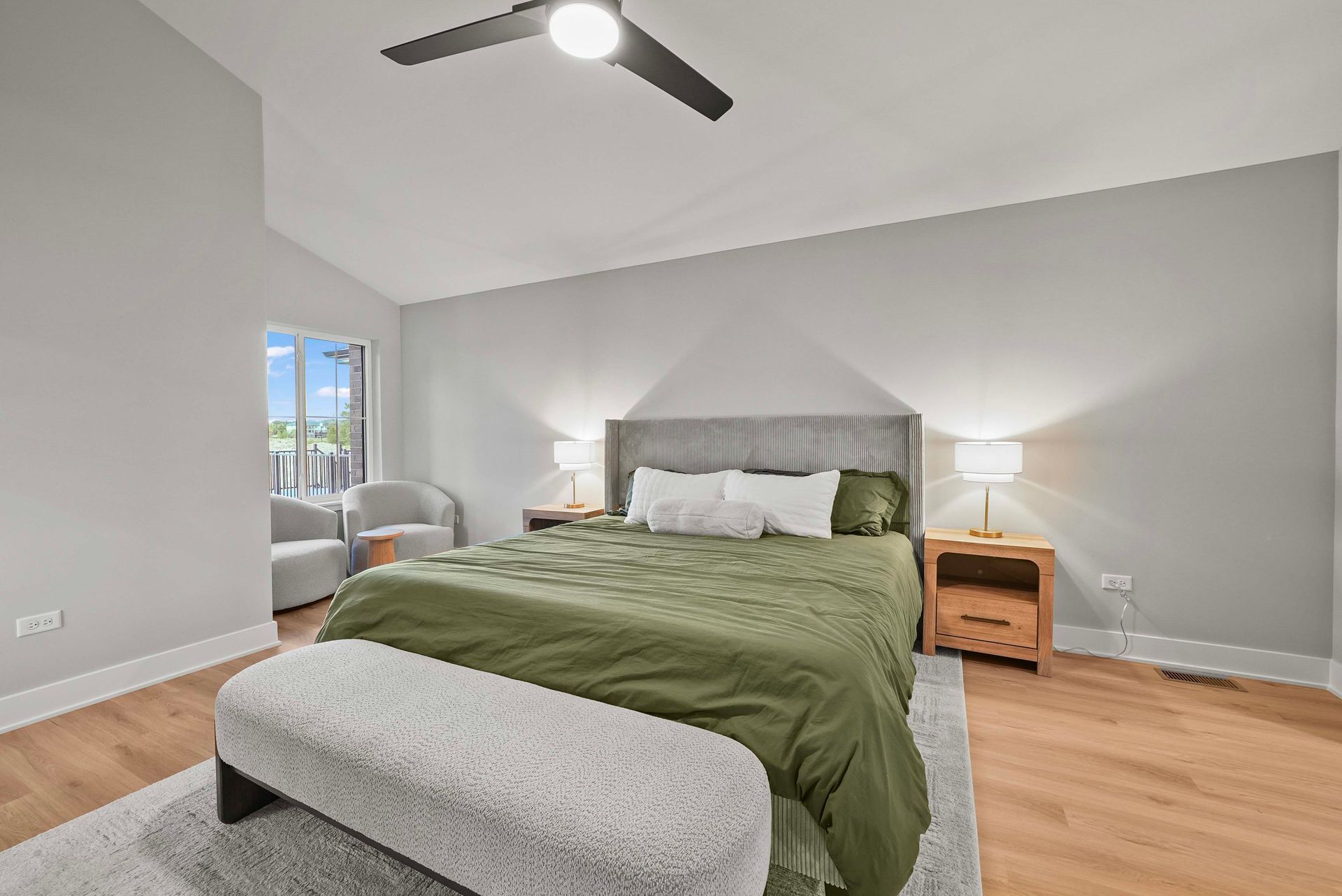 Bright bedroom with a bed, green comforter, gray walls, ceiling fan, and bedside lamp; window seating at left.